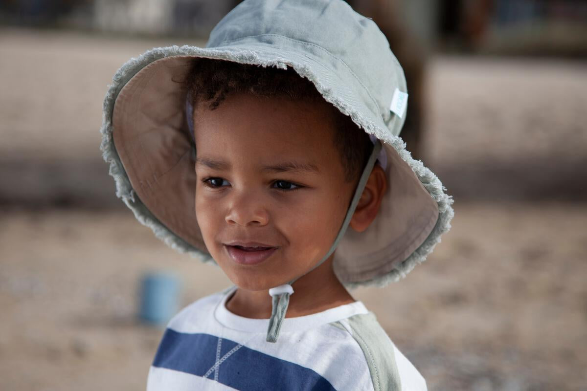 A young child with curly hair smiles outdoors on sand, wearing the ACORN KIDS Frayed Bucket Hat Khaki and a striped shirt.