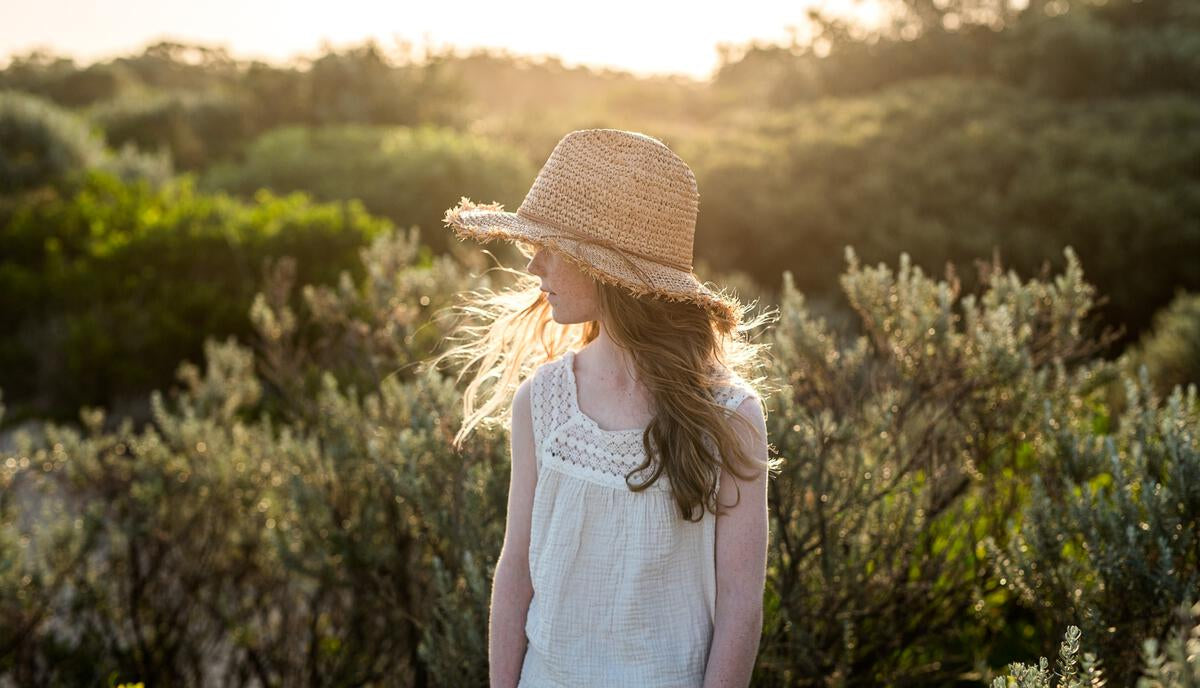 A young girl wears the ACORN KIDS Coco Straw Hat and a white sleeveless top, standing outdoors in soft sunlight among green shrubs, looking to the side.