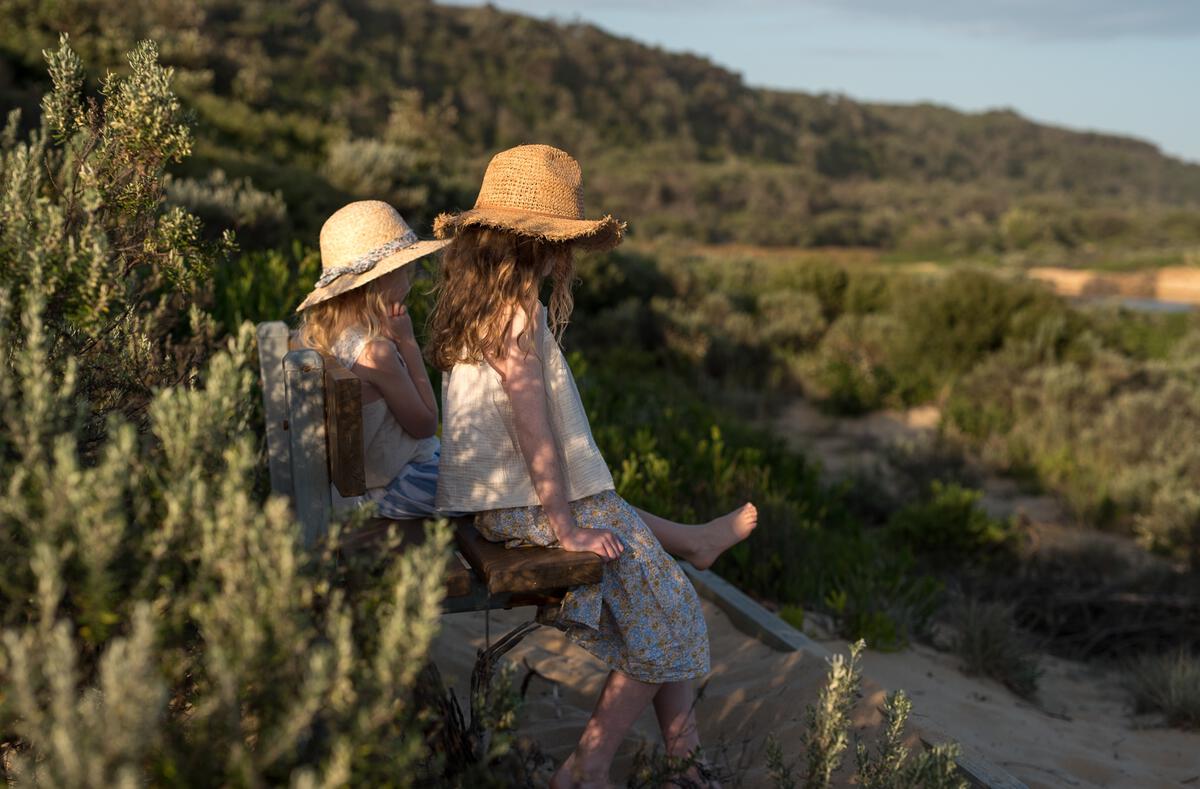 Two young girls wearing ACORN KIDS Acorn Kids - Coco Straw Hats sit on a wooden bench, surrounded by green bushes and gazing at a sunlit landscape with hills in the background.