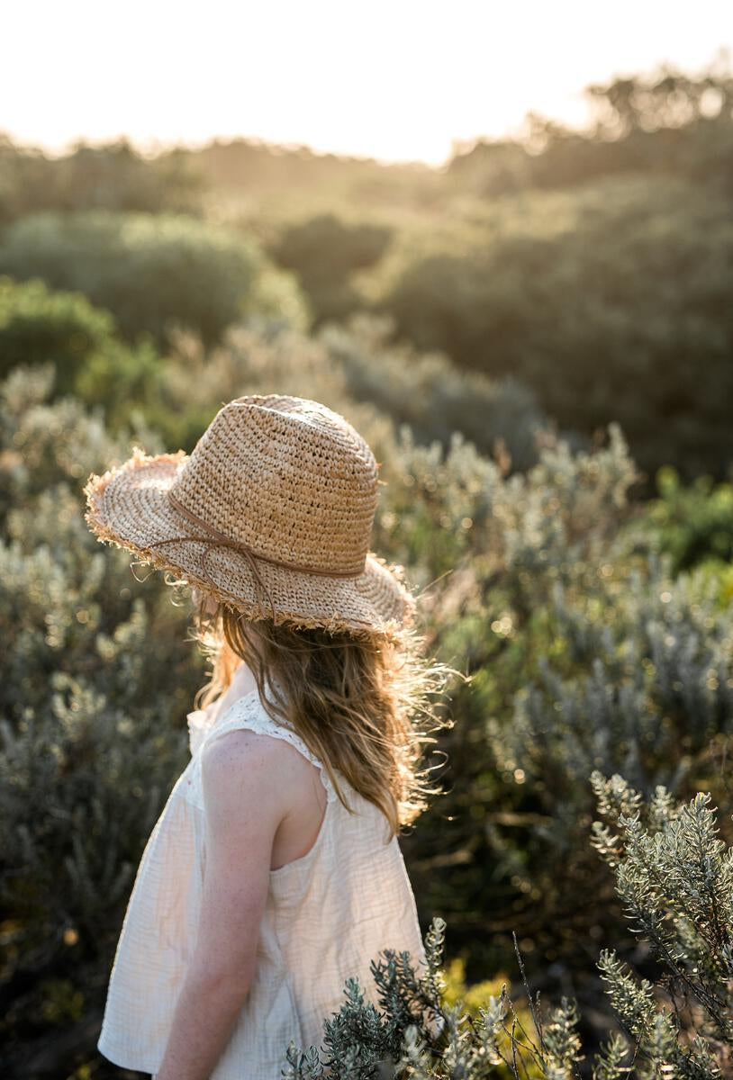 A young girl in the Acorn Kids - Coco Straw Hat by ACORN KIDS stands in a sunlit field, facing the bright horizon and enjoying stylish sun protection.