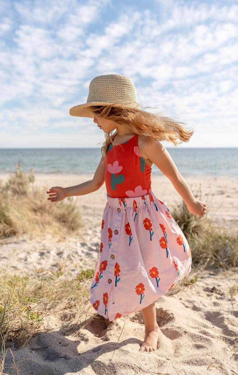 A young girl wearing the ACORN KIDS Poet Crochet Straw Hat twirls on a sandy beach in a bright floral dress, with the blue sky, scattered clouds, sea, and grassy dunes in the background.