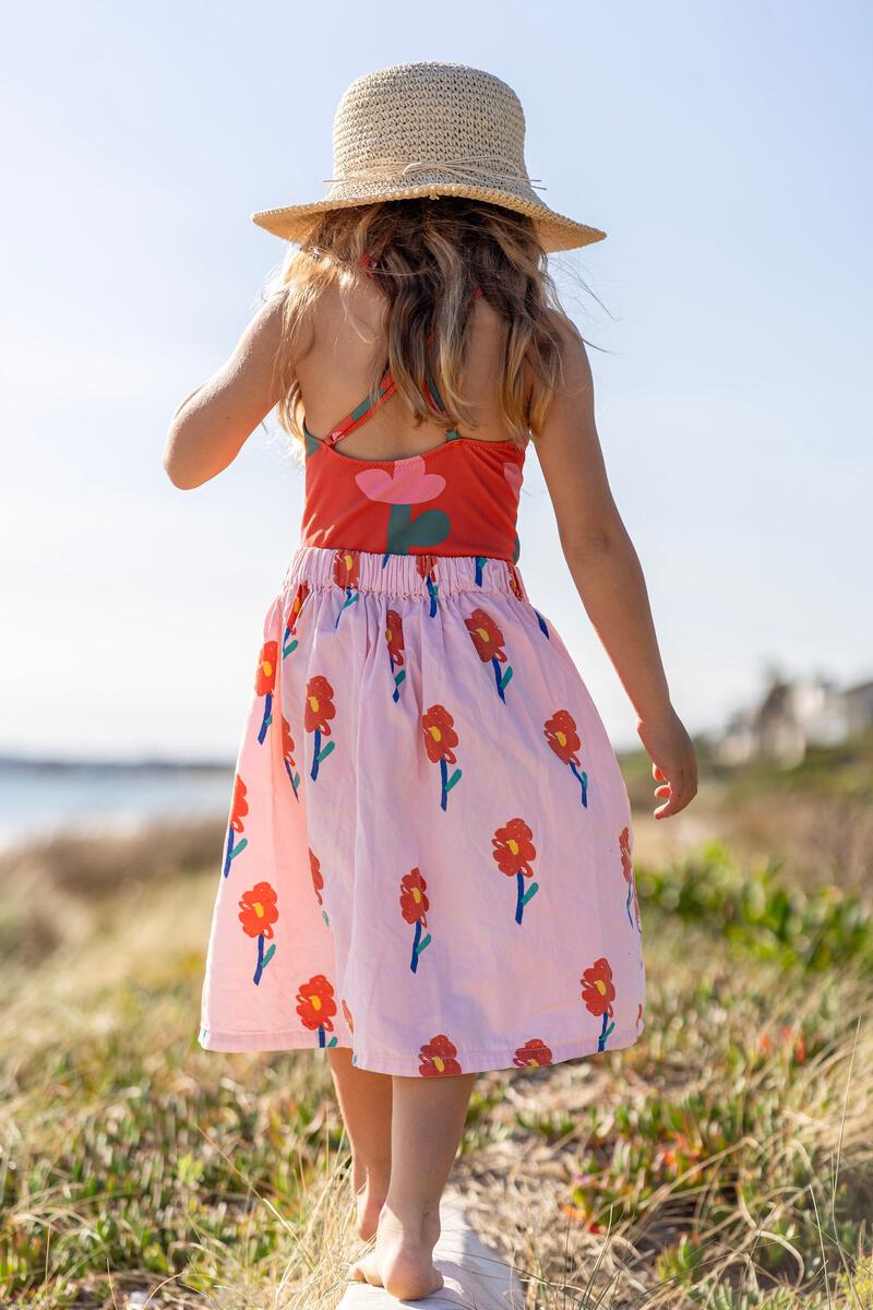 A young girl in a colorful dress with red flowers walks barefoot along a grassy beach path under a sunny sky, wearing the Acorn Kids - Poet Crochet Straw Hat by ACORN KIDS.