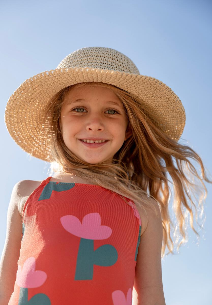 A young girl with long blonde hair smiles at the camera, wearing the ACORN KIDS Poet Crochet Straw Hat and a red swimsuit with pink and green floral patterns, standing outdoors under a clear blue sky.