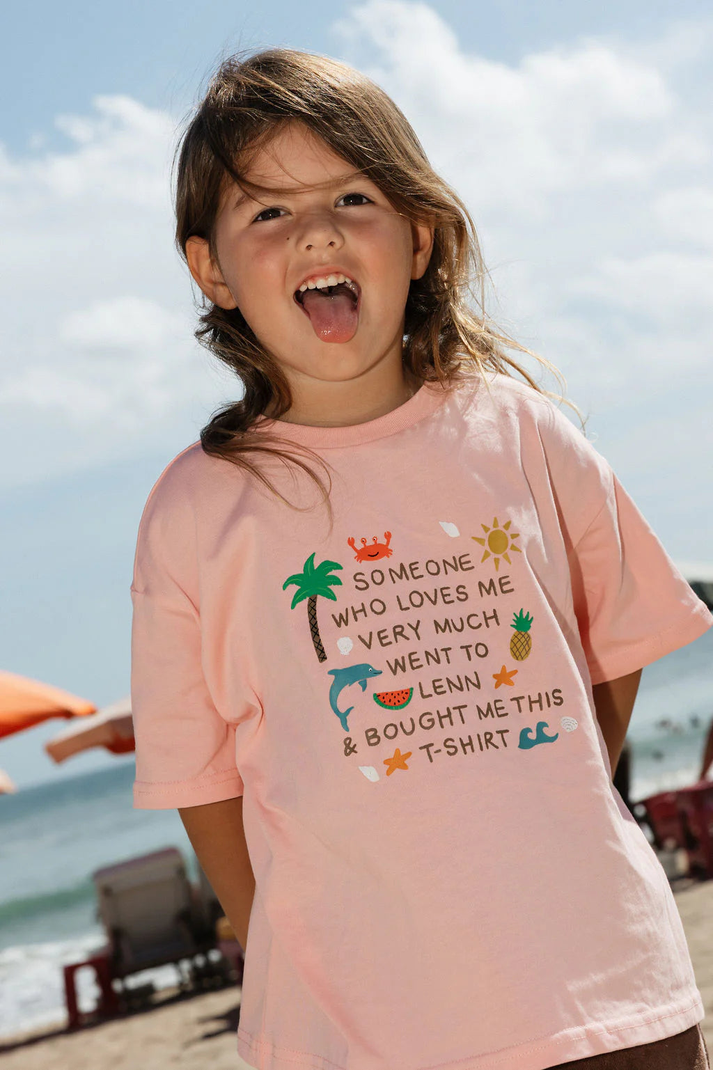 A young child smiles on a sunny beach, wearing the Lenn Label - Lenn Tee Blush by LENN LABEL. The pink 100% cotton kids tee reads, "Someone who loves me very much went to Lenny & bought me this T-shirt." Orange umbrellas and the sea are behind.