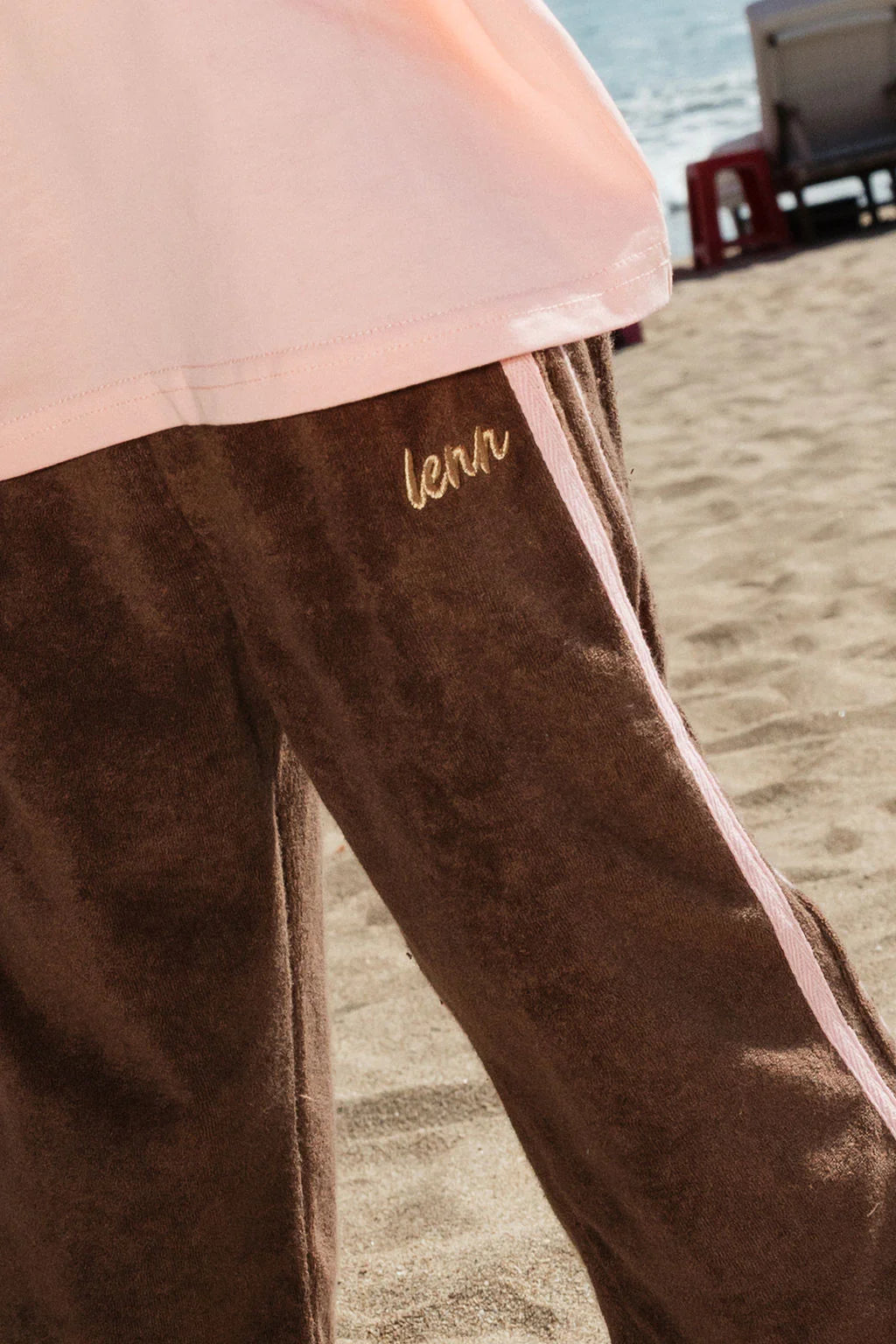 A person wearing LENN LABEL's Lenn Label - Weekend Terry Pant Choc—relaxed fit brown pants with gold "leav" embroidery and an elastic waistband—stands on a sandy beach with the ocean and beach chairs in the background.