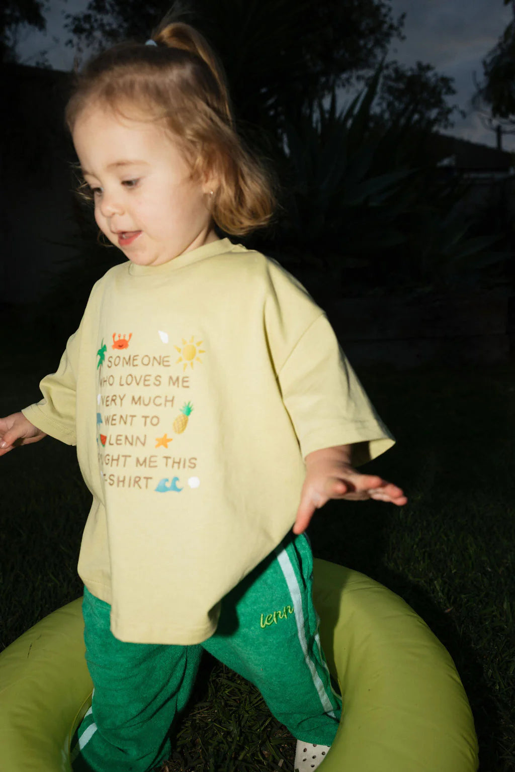 A toddler with light brown hair, wearing a relaxed fit yellow shirt and LENN LABEL’s Weekend Terry Pant Jungle stands on grass inside a green inflatable ring outdoors during the evening.