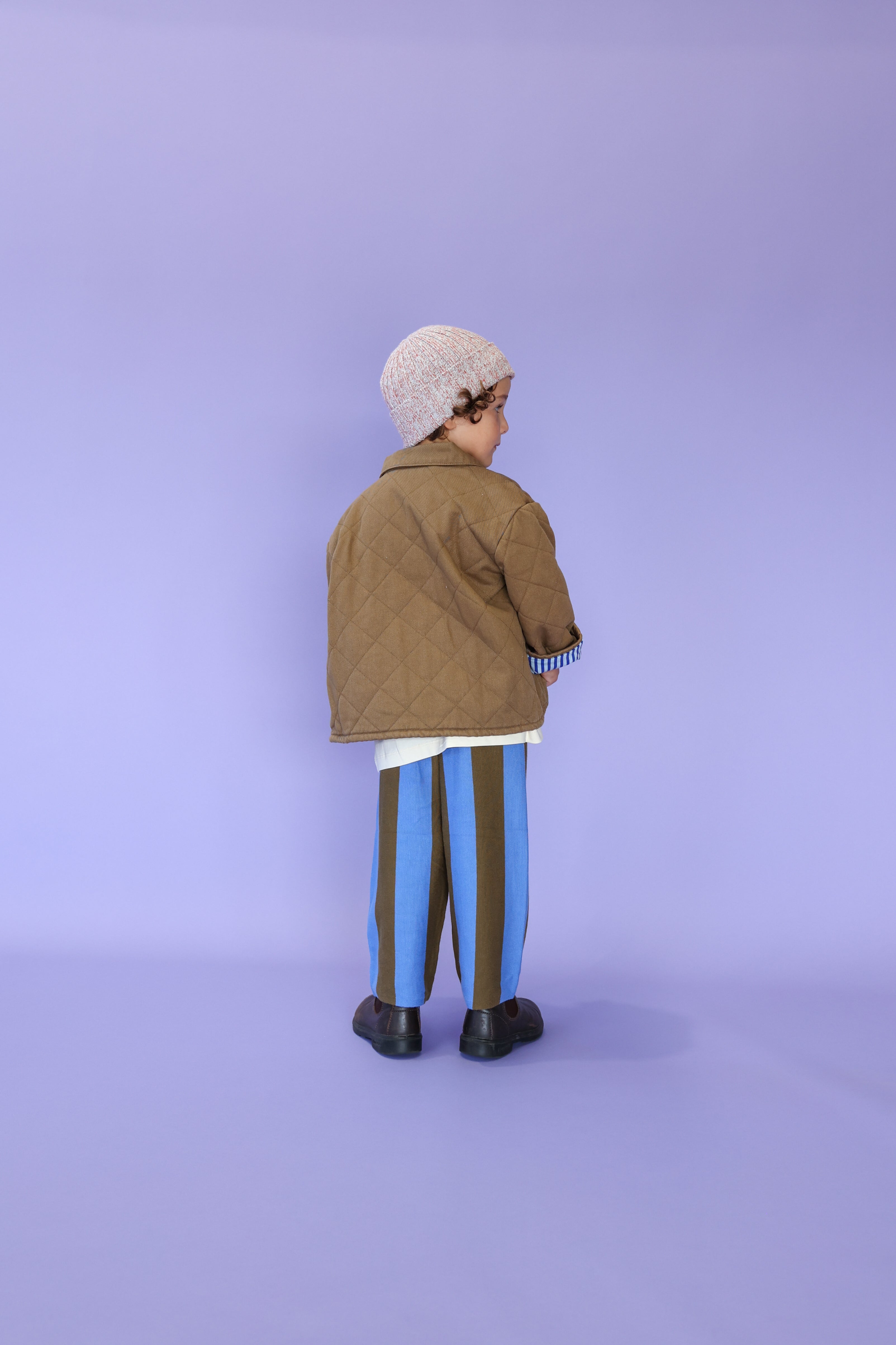 A young child stands facing away against a light purple background, wearing a brown quilted jacket, striped pants, black shoes, and the GROWN - Twisted Pixie Beanie.
