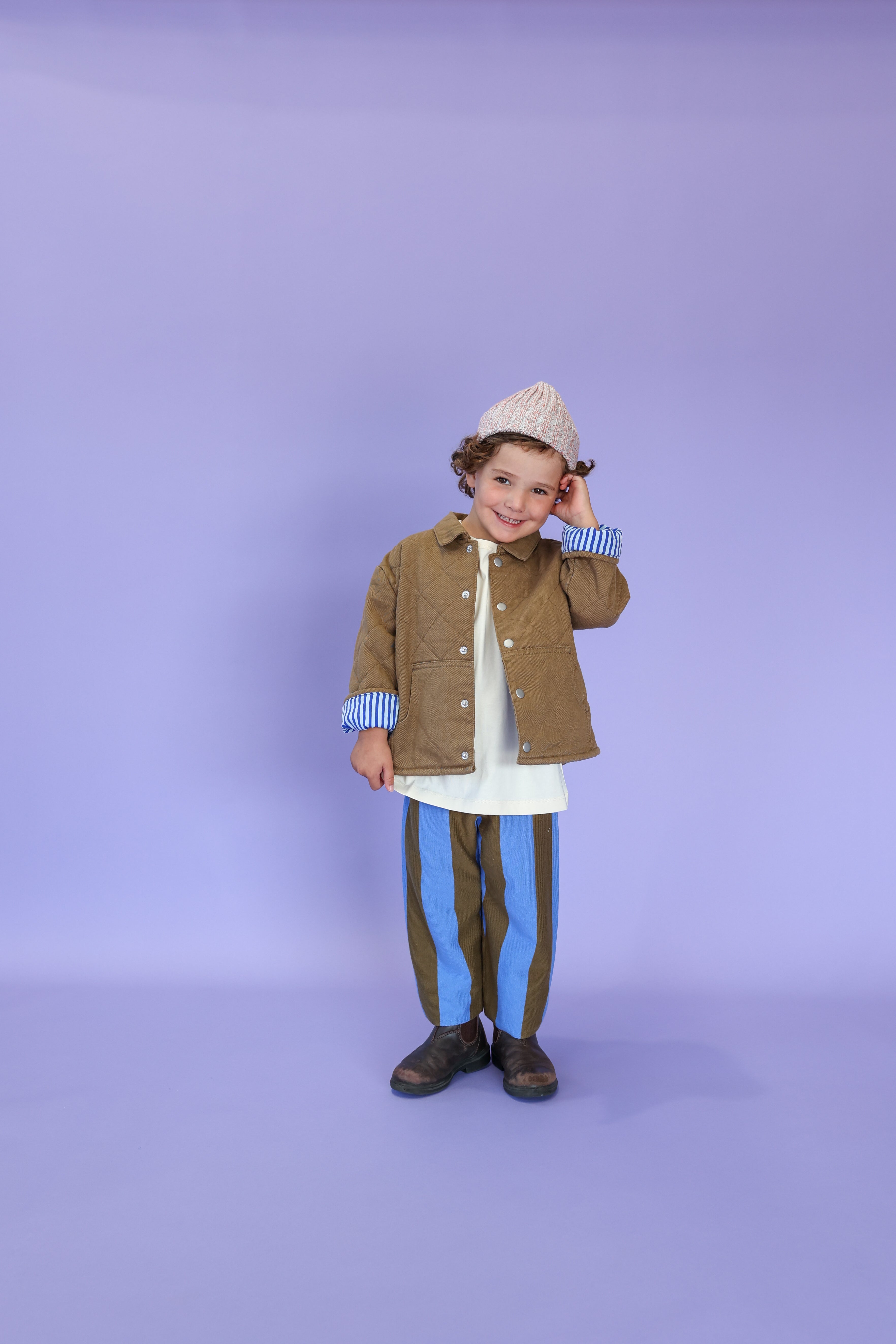 A young child smiles against a lavender background, wearing the Grown - Twisted Pixie Beanie by GROWN, a beige quilted jacket made from recycled polyester, striped blue and olive pants, and dark shoes. The child touches their hat with one hand.