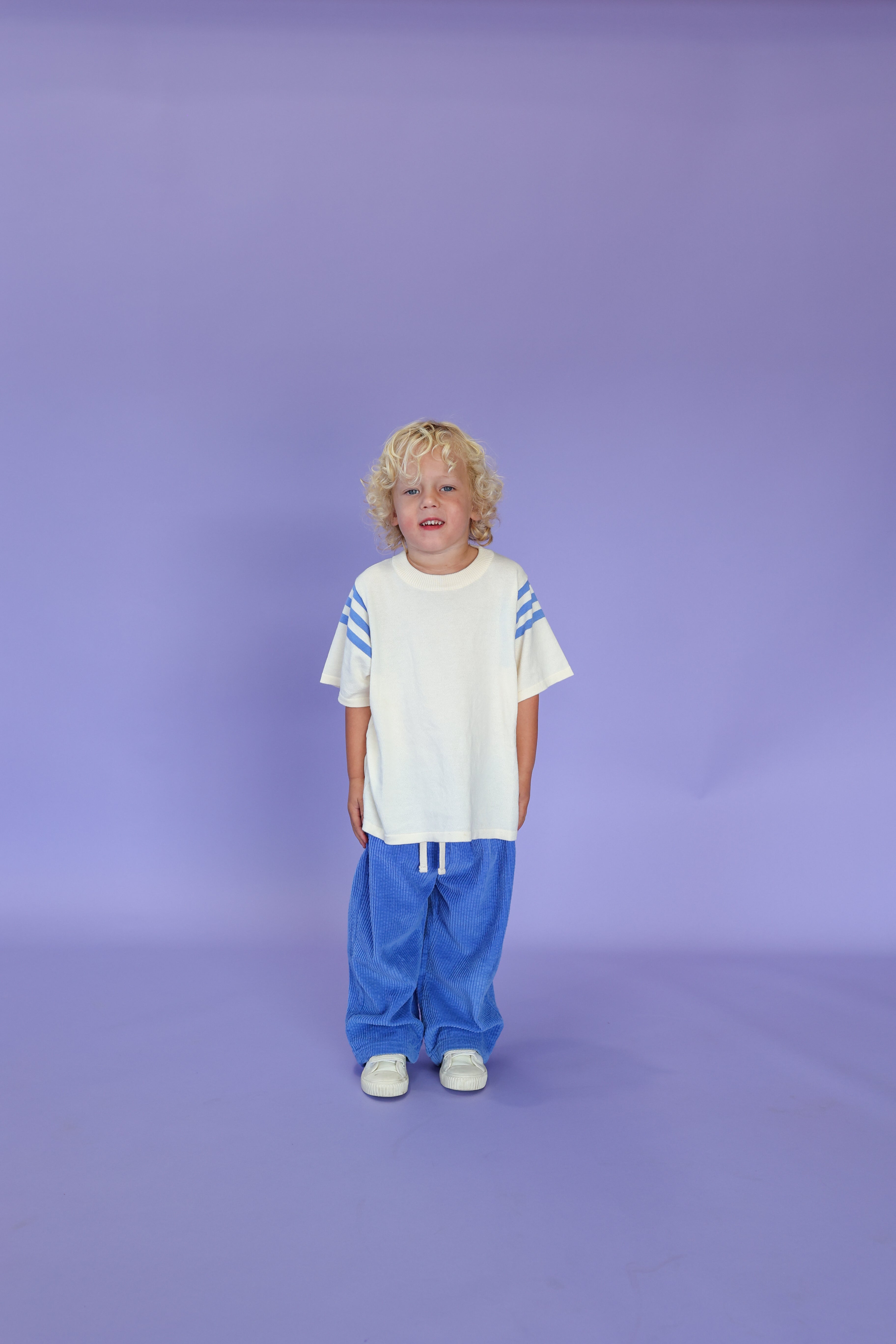 A young child with curly blond hair stands against a plain light purple background, wearing GROWNβs Organic Relaxed Cord Pant in Sea, paired with a white shirt with blue striped sleeves and white shoes.