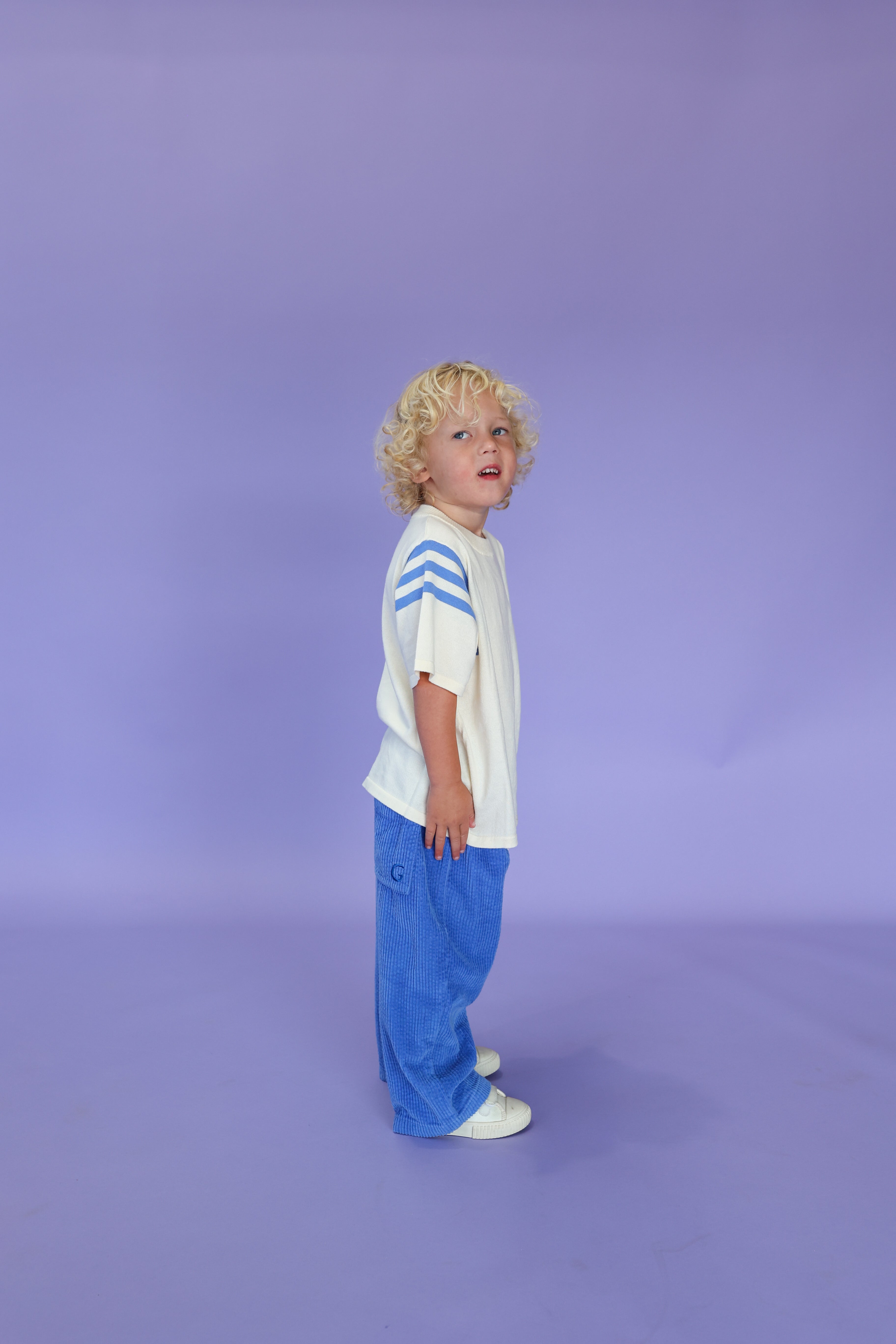 A young child with curly blond hair stands sideways on a light purple background, wearing GROWN's Organic Relaxed Cord Pant in Sea, paired with a relaxed fit white shirt with blue striped sleeves and white shoes.