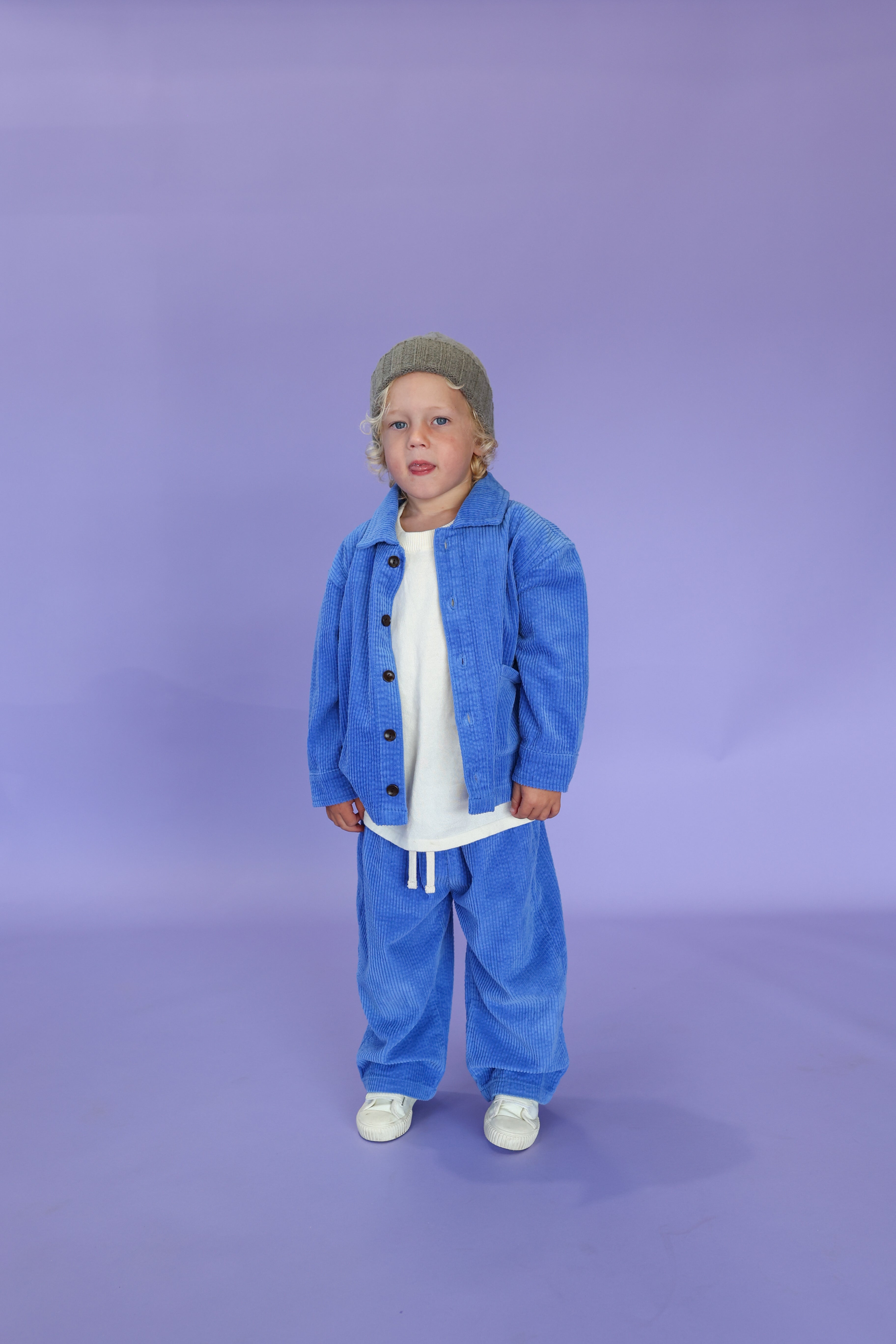 A young child wears GROWN's Organic Relaxed Cord Pant in Sea, paired with a gray beanie, blue button-up jacket, white t-shirt, and white shoes, standing against a plain light purple background.