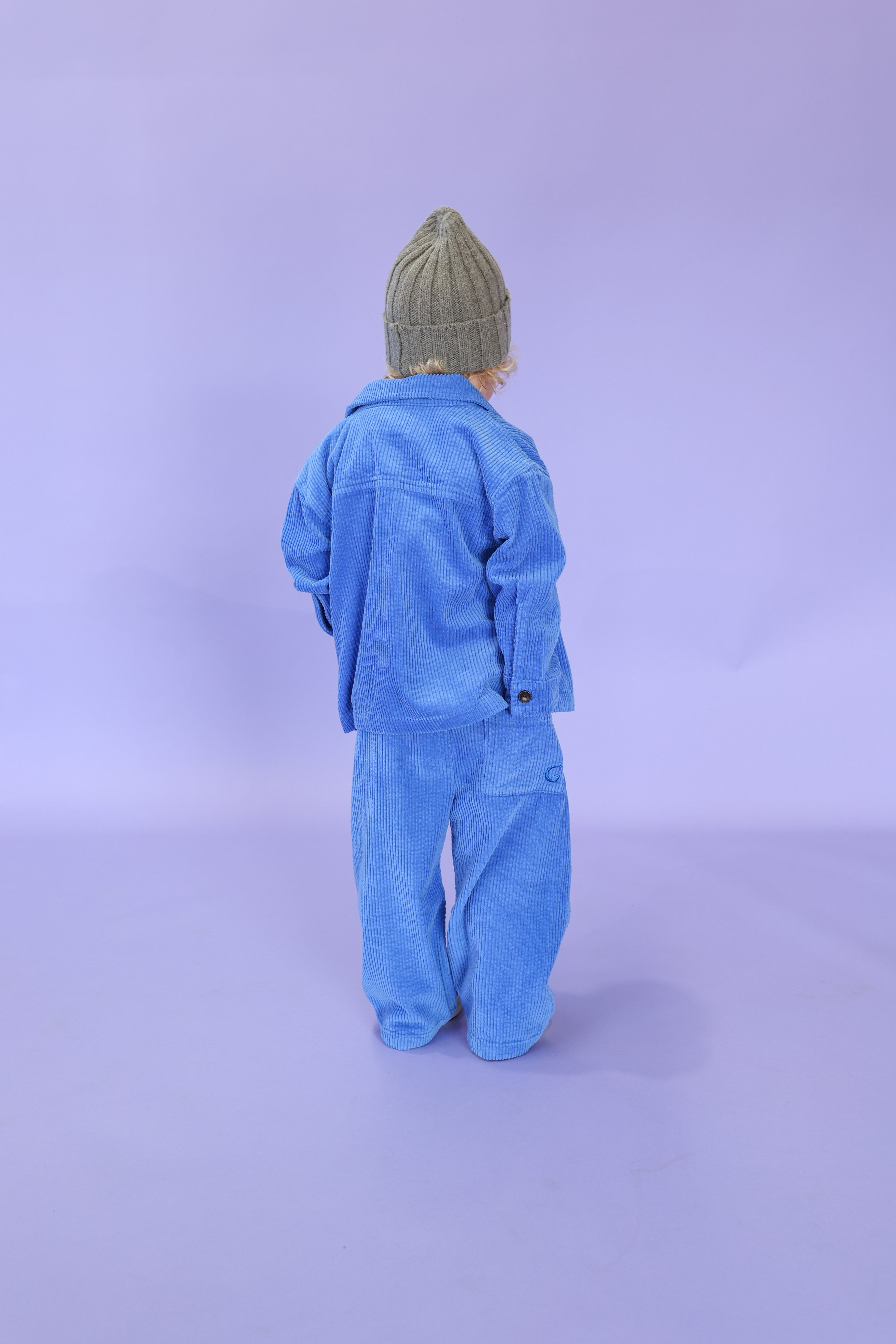 A child wearing a gray knit beanie and GROWN Organic Relaxed Cord Pant Sea stands facing away against a light purple background.