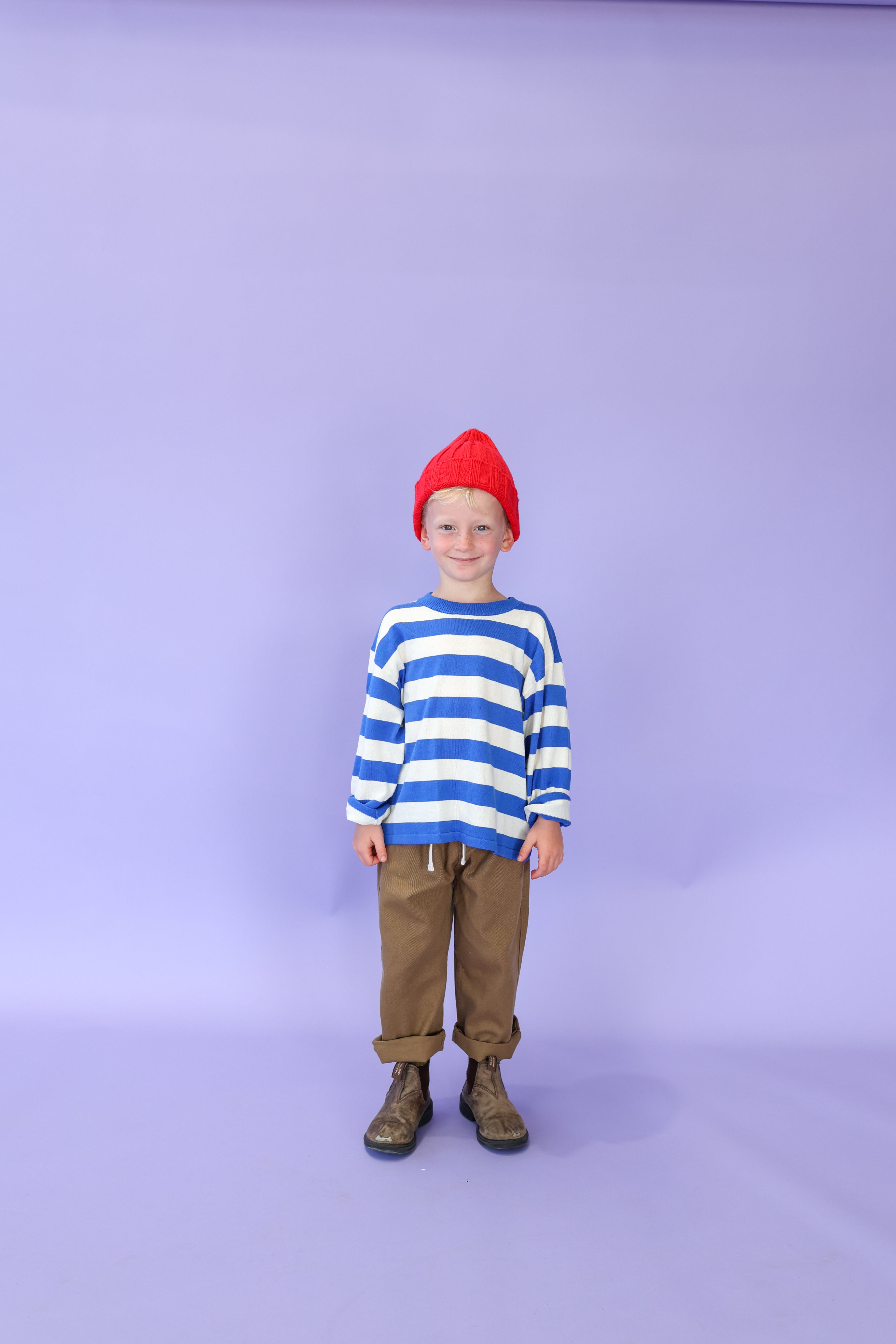 A young child wears the GROWN - Merino Blend Ribbed Beanie Flame by GROWN, smiling against a plain light purple background.