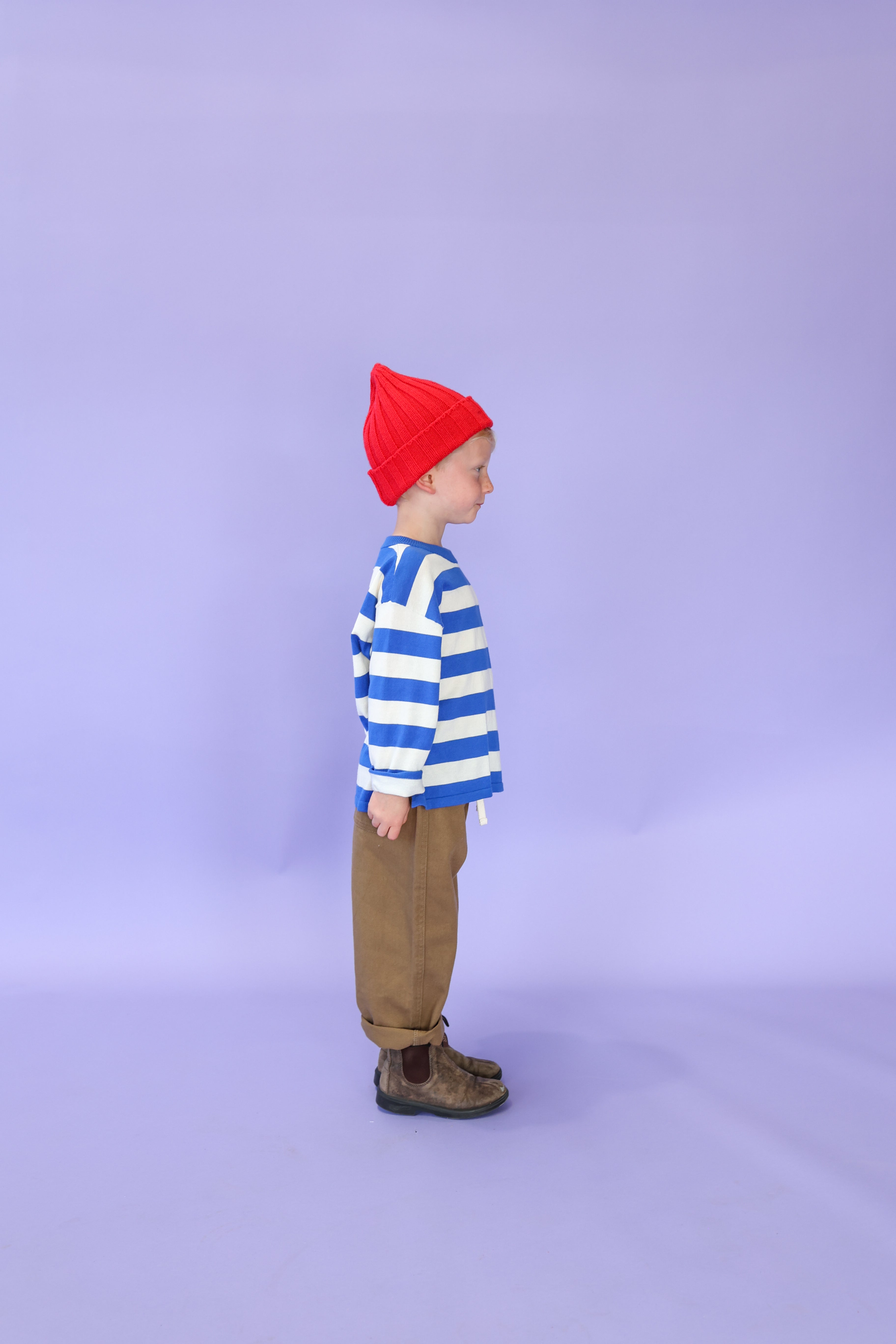 A young child stands in profile against a light purple background, wearing the GROWN Merino Blend Ribbed Beanie Flame in red, a blue-and-white striped shirt with anchor details, brown pants, and brown boots.