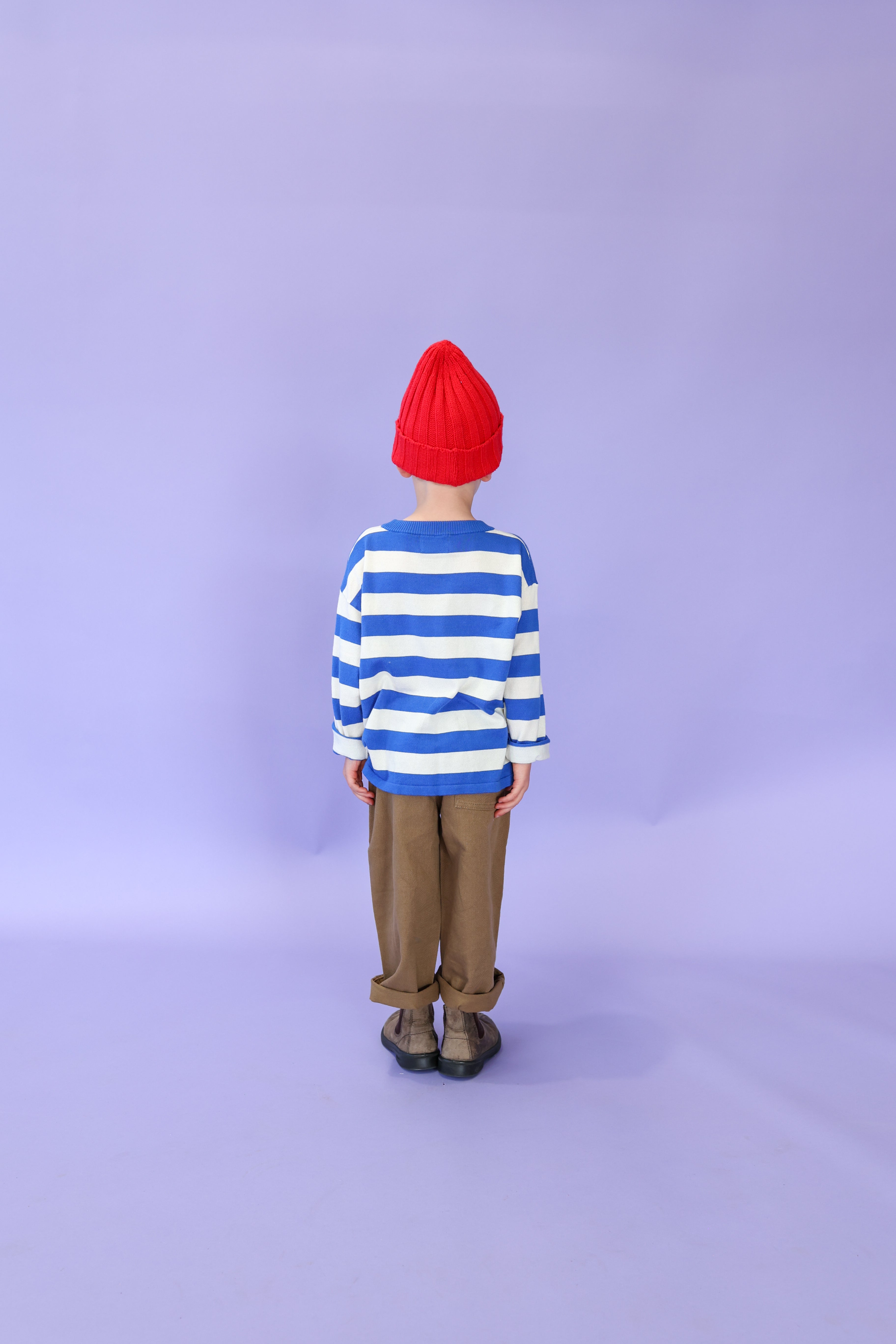 A child faces away, wearing the GROWN Merino Blend Ribbed Beanie Flame in red, a blue-and-white striped organic cotton sweater, brown pants, and black shoes against a plain light purple background.