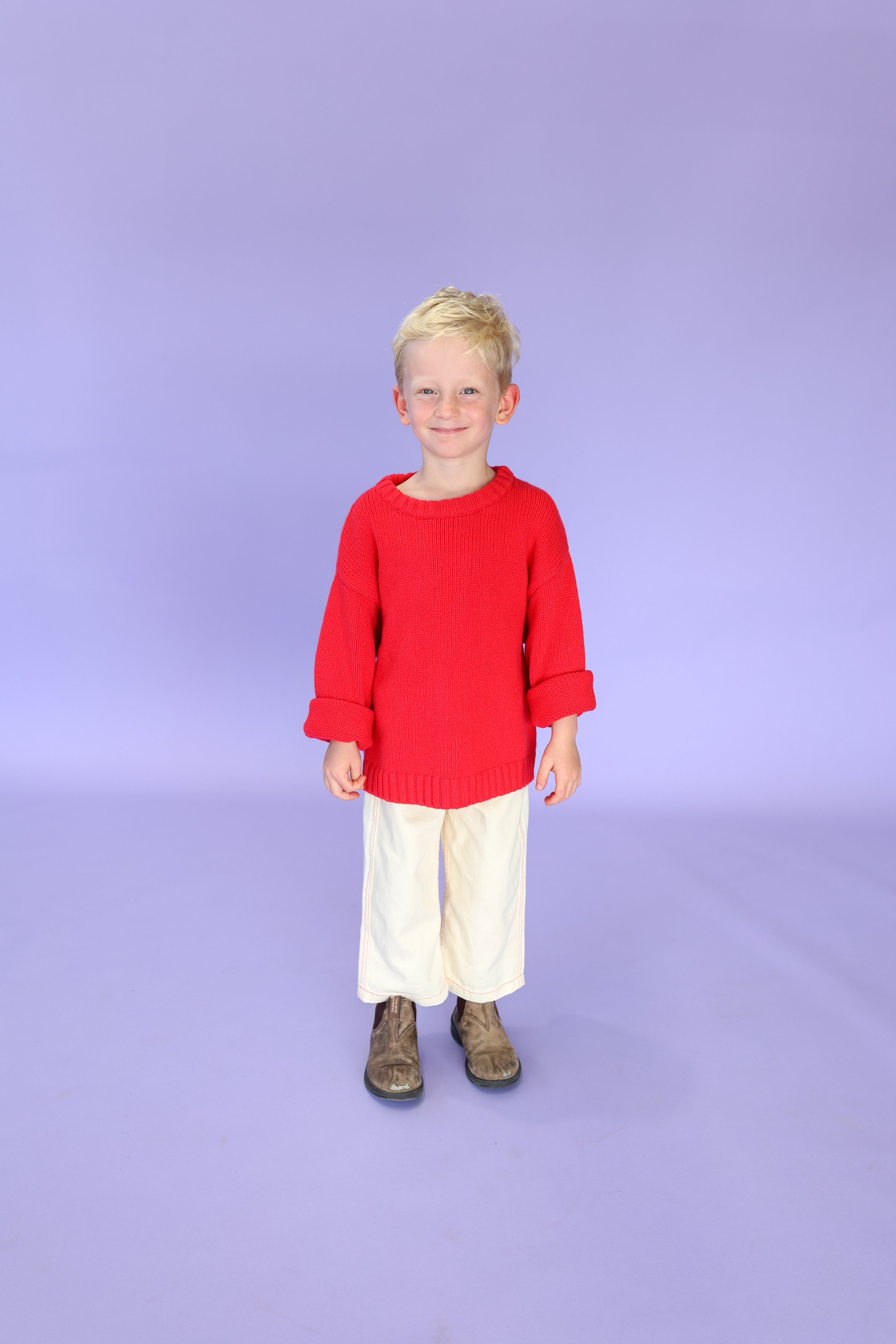 A young child with short blond hair smiles against a light purple background, wearing the GROWN Merino Blend Everyday Pull Over Flame sweater, off-white pants, and brown boots.