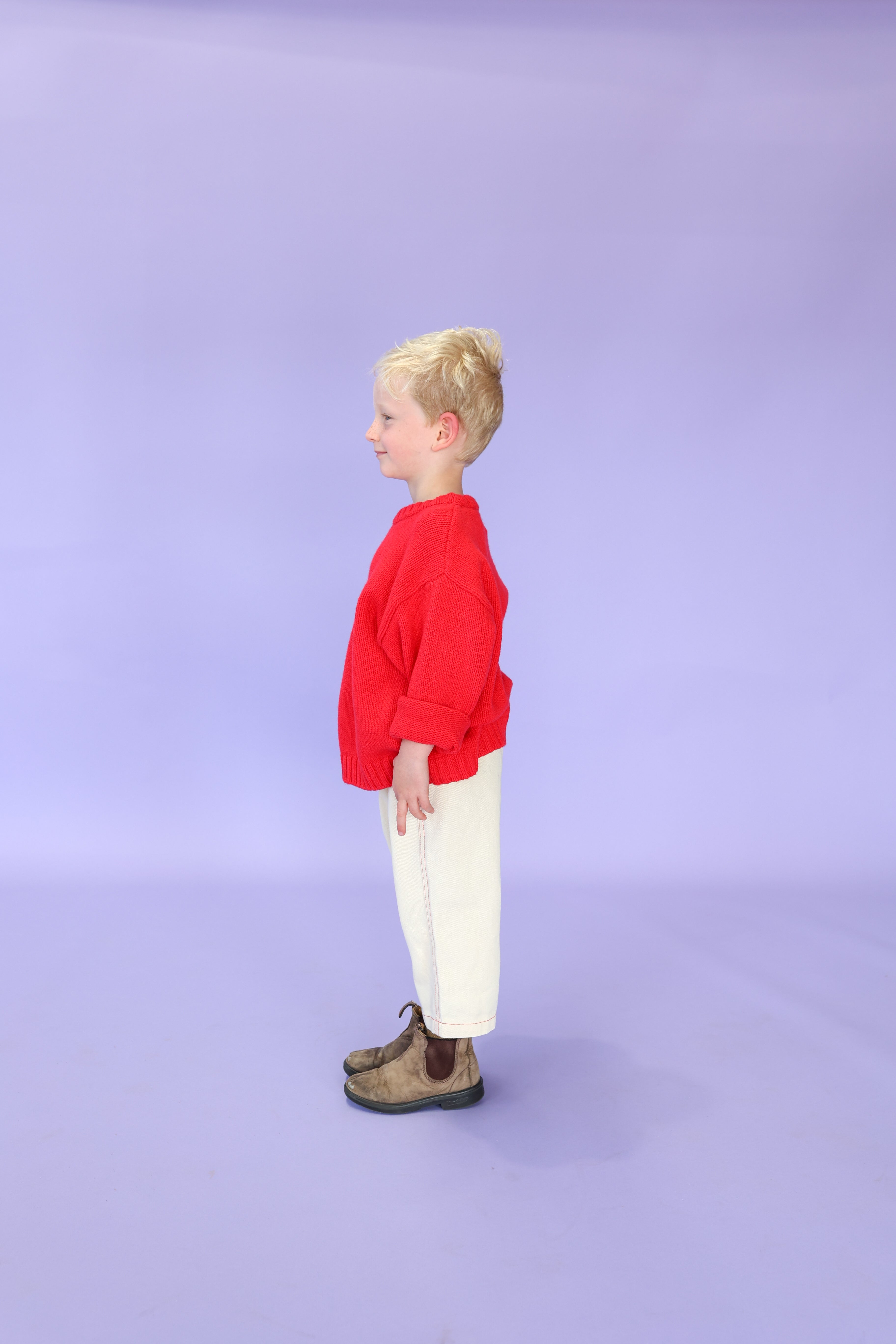 A young child with blond hair stands in profile against a plain lavender background, wearing the GROWN Merino Blend Everyday Pull Over Flame red sweater by GROWN, paired with white pants and brown shoes.