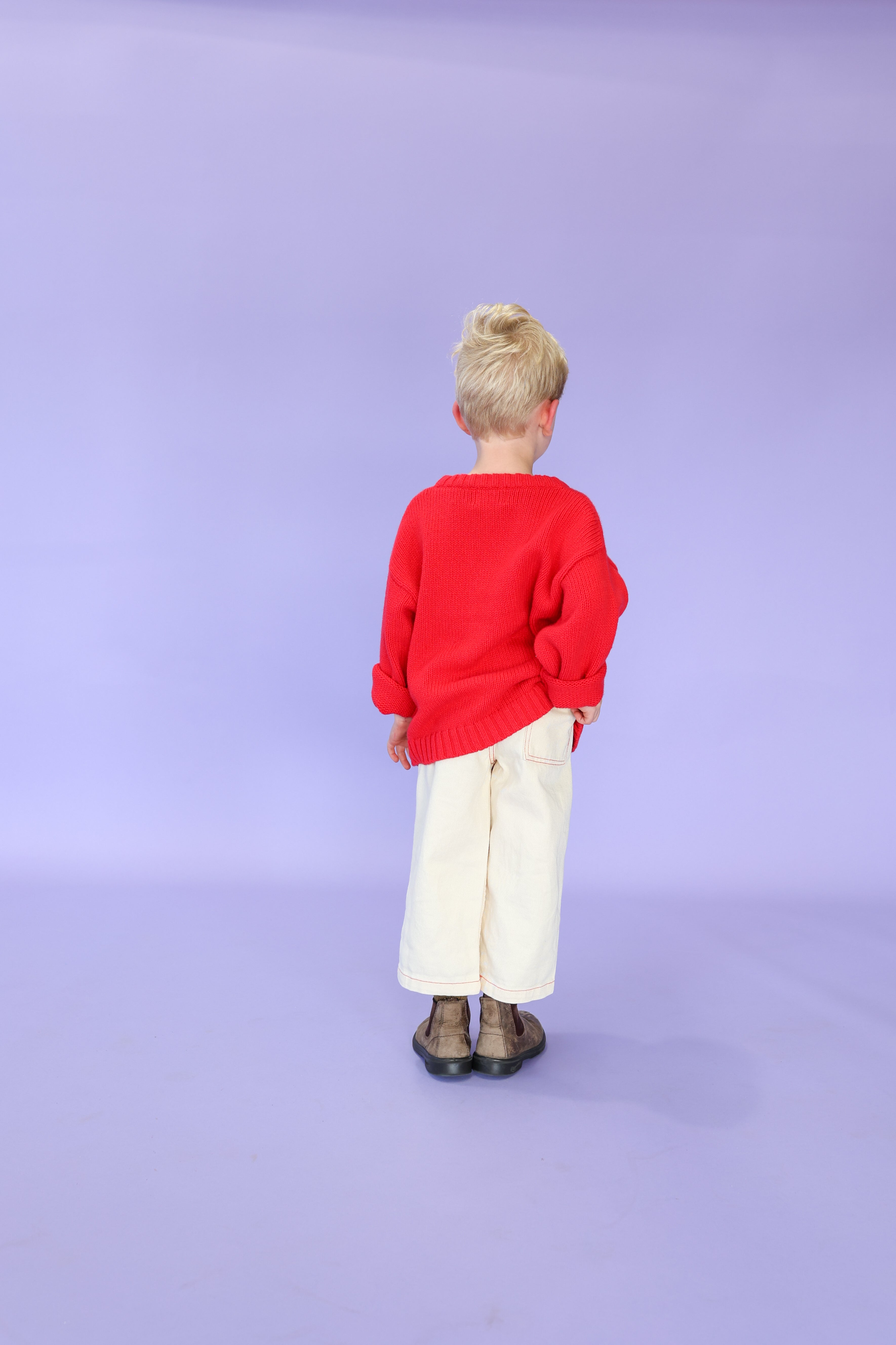 A young child with blond hair, wearing the GROWN Merino Blend Everyday Pull Over in Flame, cream-colored pants, and brown shoes stands facing away against a light purple background.