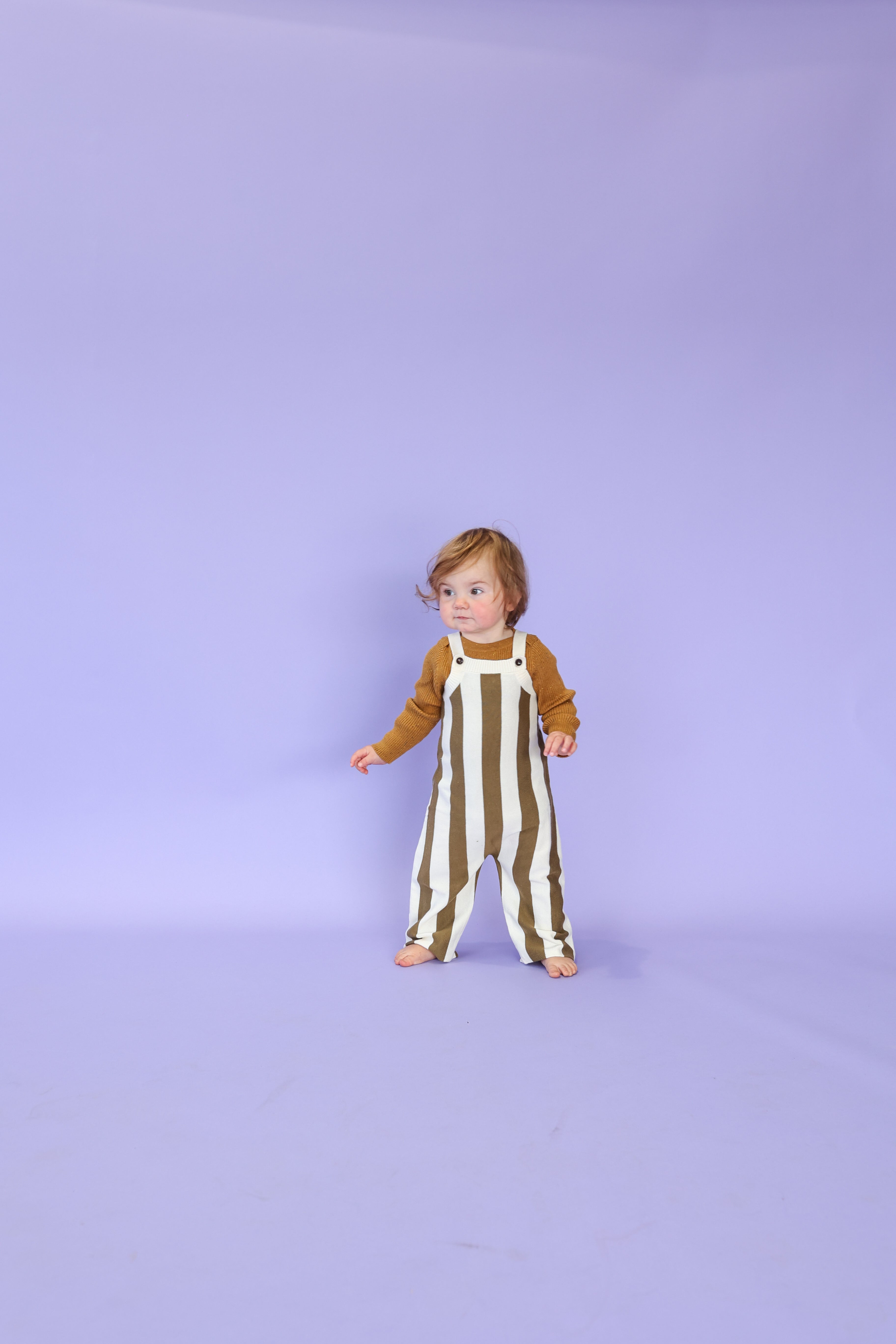 A toddler stands barefoot against a plain lavender background, wearing the GROWN Vertical Stripe Knit Overallsâwhite organic cotton with thick brown stripes and adjustable strapsâpaired with a brown long-sleeve shirt.