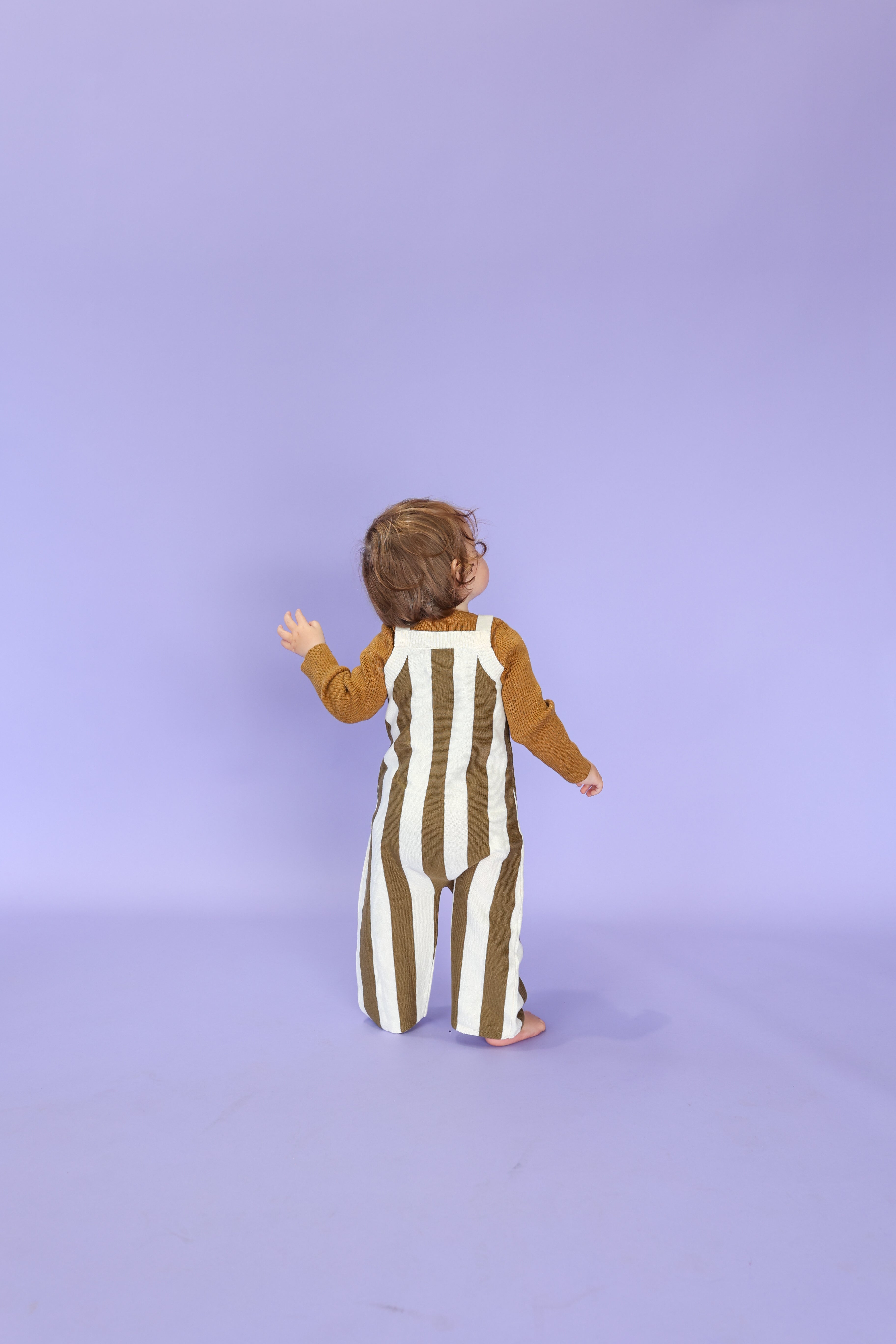 A toddler with brown hair, wearing GROWN's Grown - Vertical Stripe Knit Overalls made from GOTS certified organic cotton, stands barefoot on a light purple floor facing a matching wall.