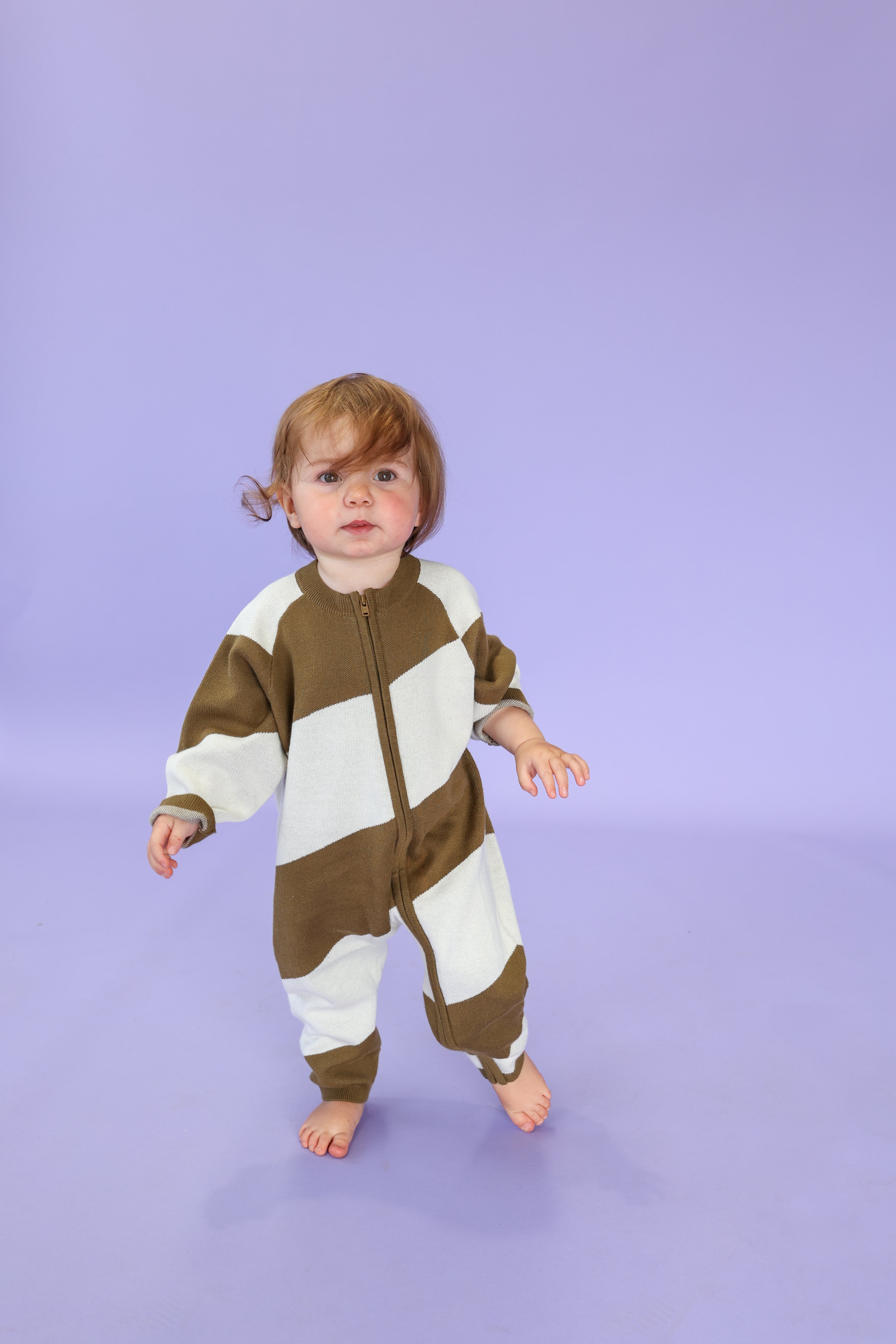 A toddler with light brown hair stands barefoot on a solid lavender floor, wearing the GROWN Organic Essential Zip Starsuit Coffee Stripe made from organic cotton.