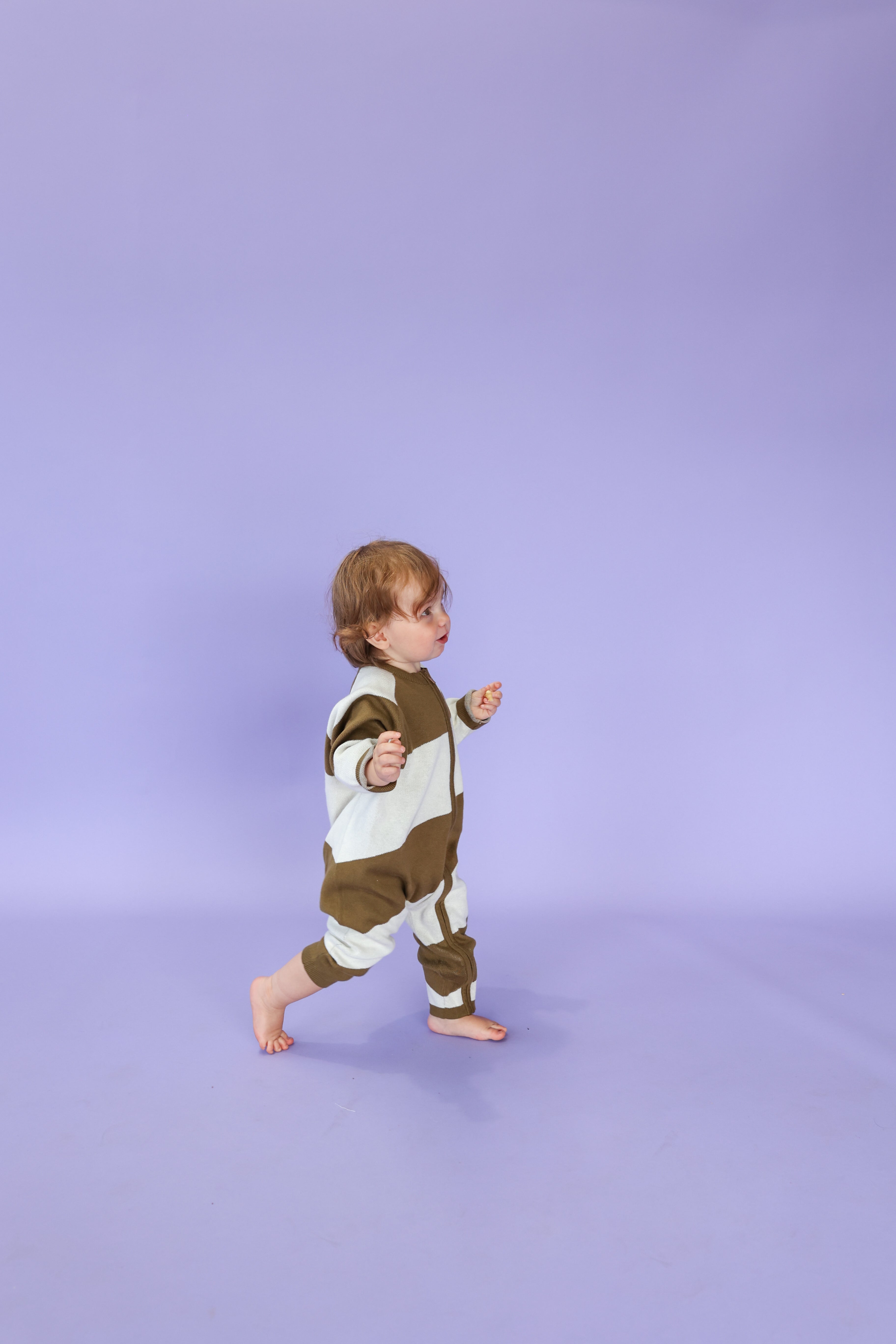 A young child wearing the GROWN Organic Essential Zip Starsuit Coffee Stripe walks barefoot across a light purple floor with a plain purple background.