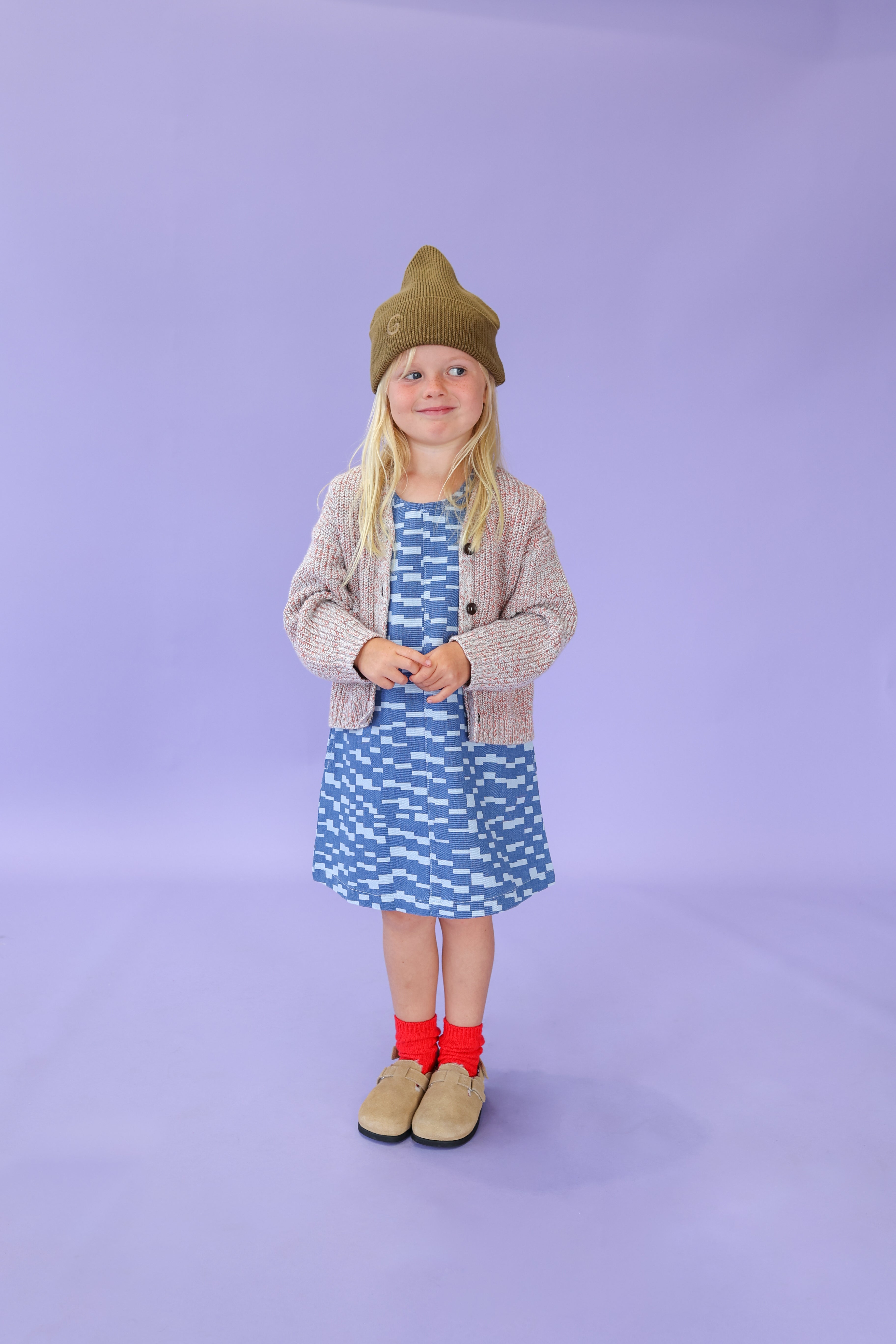 A young child with long blonde hair wears the GROWN - Twisted Chunky Rib Cardigan, a green chunky rib knit beanie, blue patterned dress, red socks, and tan shoes, smiling against a light purple background.