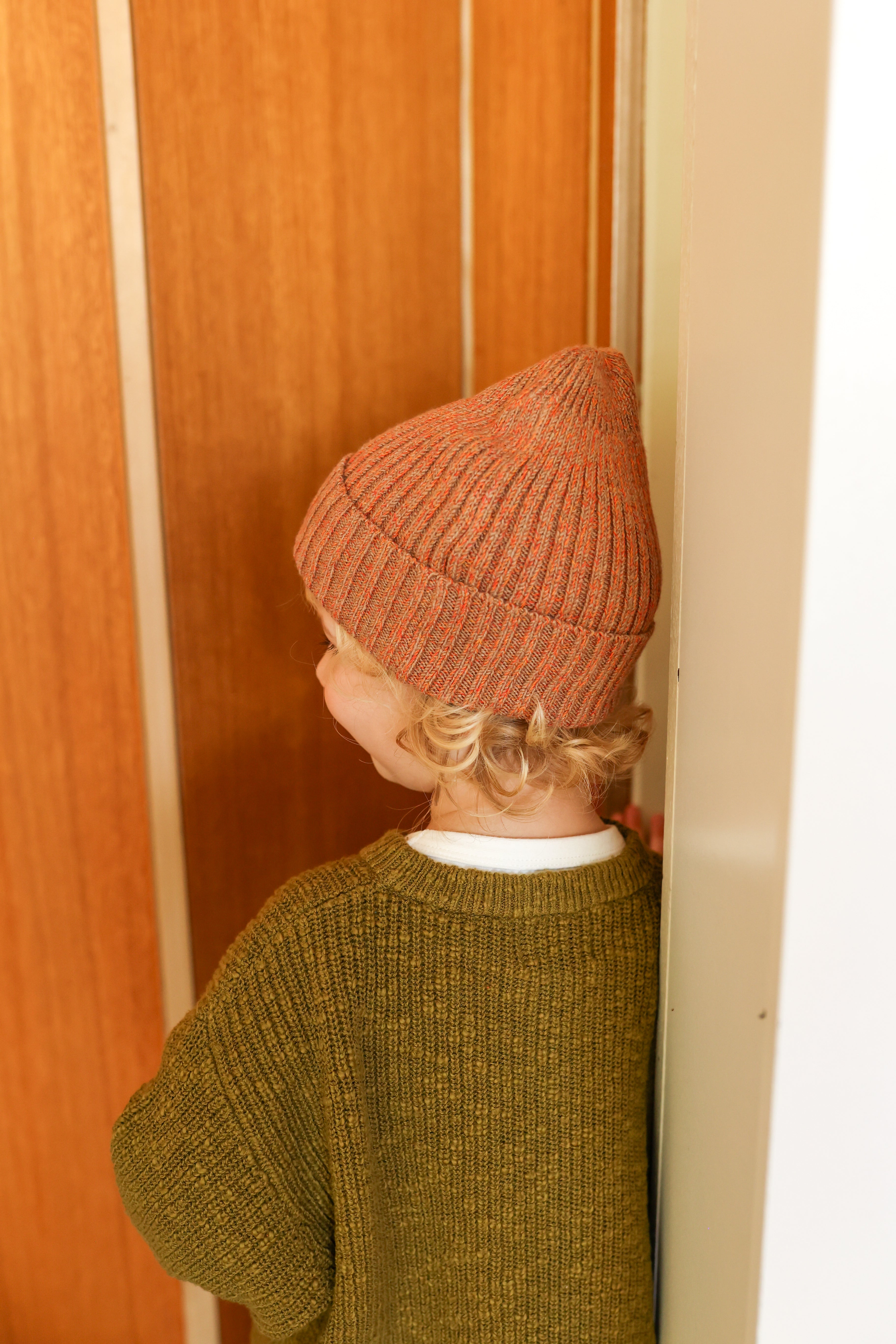 A child with curly hair, wearing a Grown ~ Fleck Yarn Pixie Beanie Tawny from GROWN and a green sweater, stands facing a wooden door. The child is partly against a white wall, softly illuminated by light.