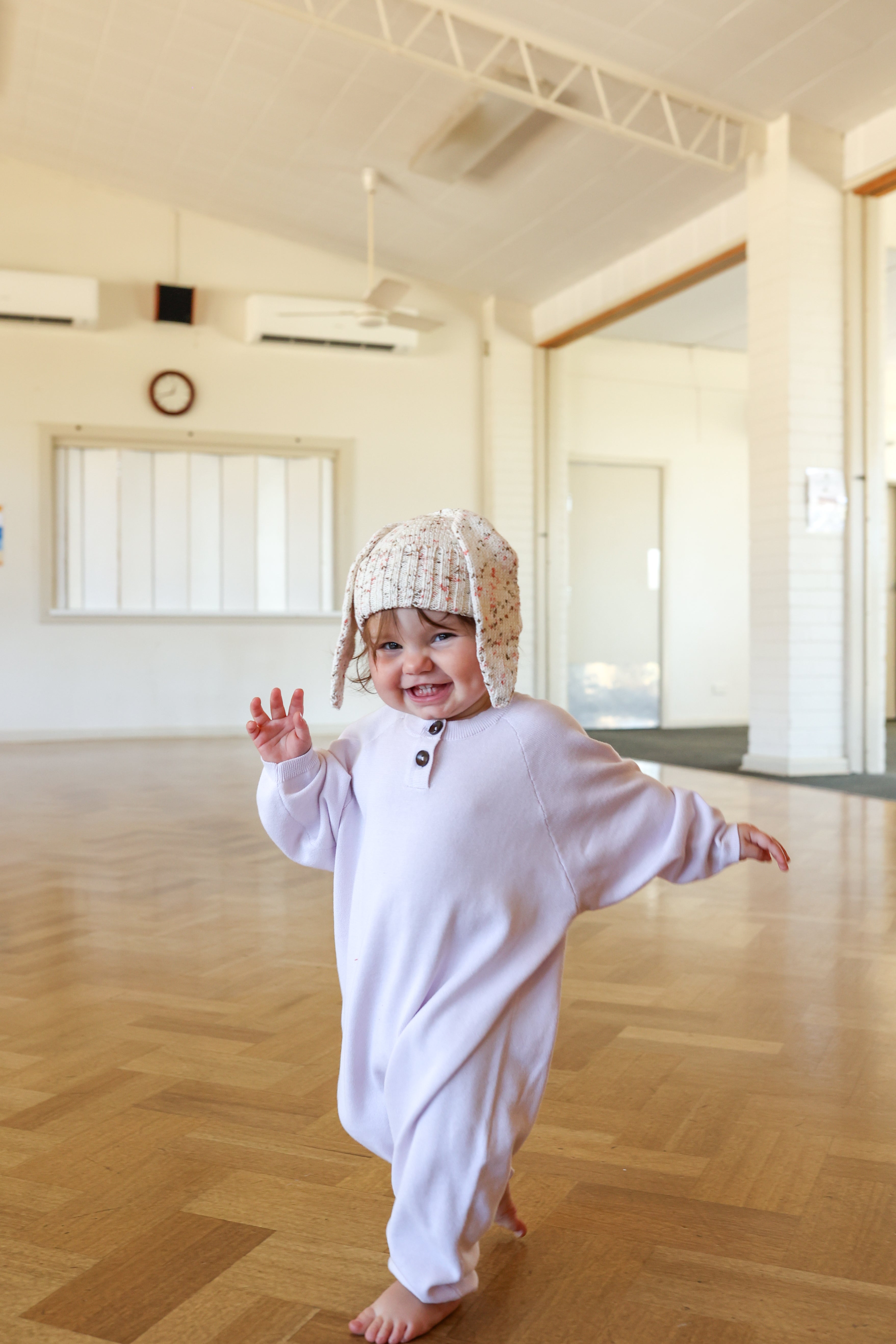 A small child in a cozy white onesie and an adorable GROWN Funfetti Bunny Beanie Pokie walks barefoot on a wooden floor indoors, smiling and waving. The spacious room features a clock and windows in the background.