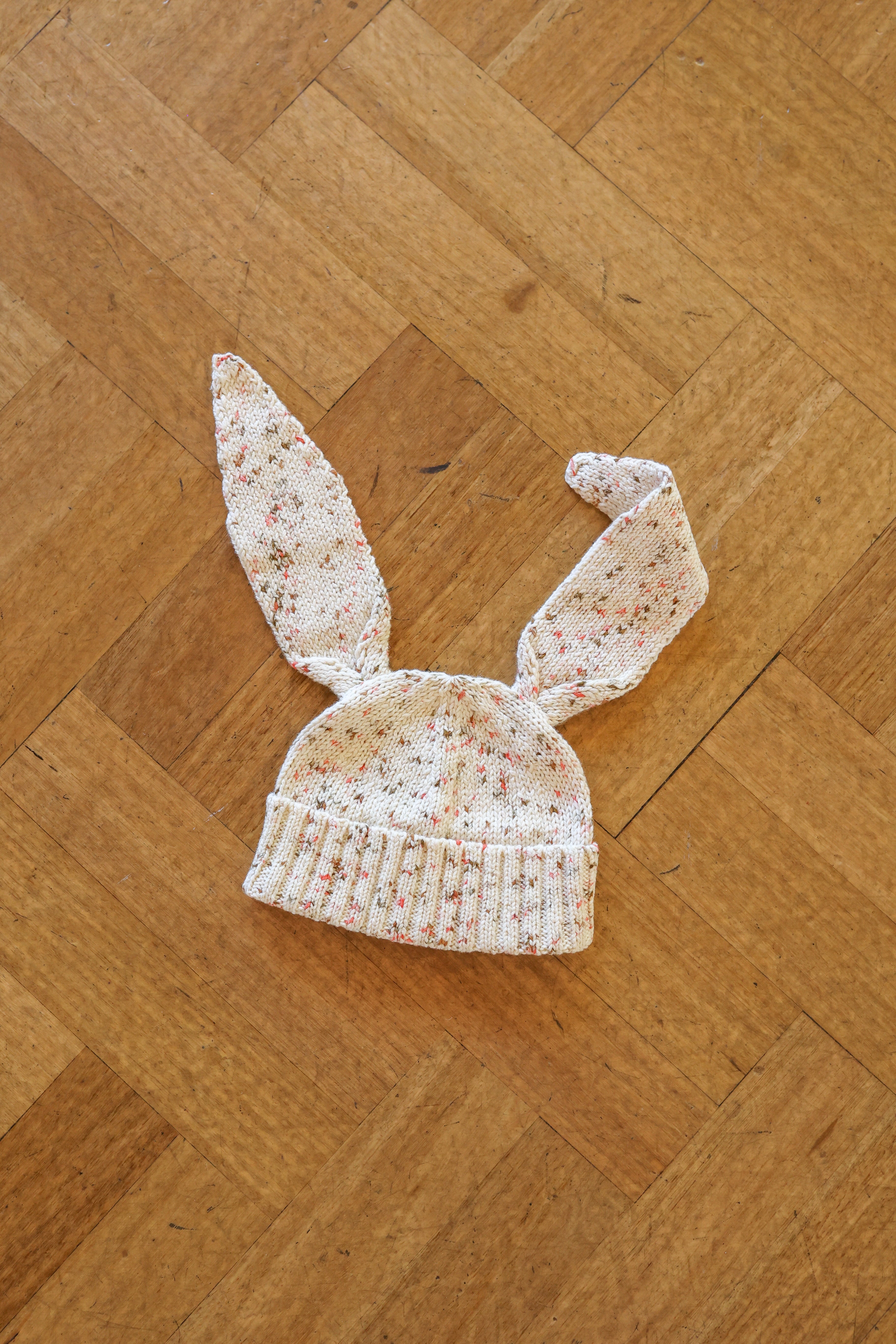 The GROWN Funfetti Bunny Beanie Pokie, a jersey knitted baby hat with long bunny ears and red speckles, rests on a wooden parquet floor. Made from organic cotton for superior comfort.