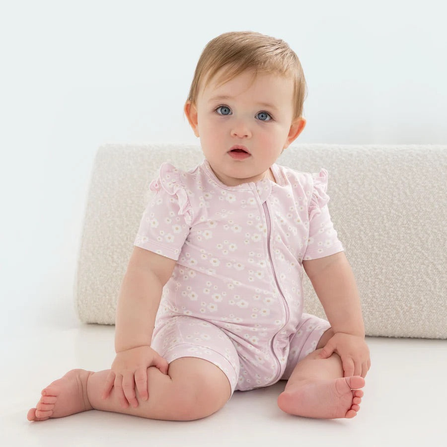 A baby in KYND BABY's Day or Night Short Sleeve Frill Onesie Orchid Bloom sits on a light surface. The soft viscose fabric features pale pink with white floral patterns. With short, light brown hair, the baby gazes at the camera neutrally, one hand resting on the floor.