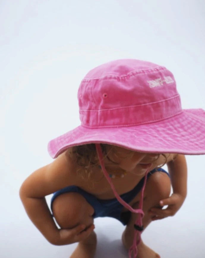 A young child squats down wearing the Ziggy Zaza - Everyday Scout Hat in Flamingo by ZIGGY ZAZA, made of cotton canvas, paired with blue shorts. The hat's adjustable chin strap hangs loosely against a plain, light background.