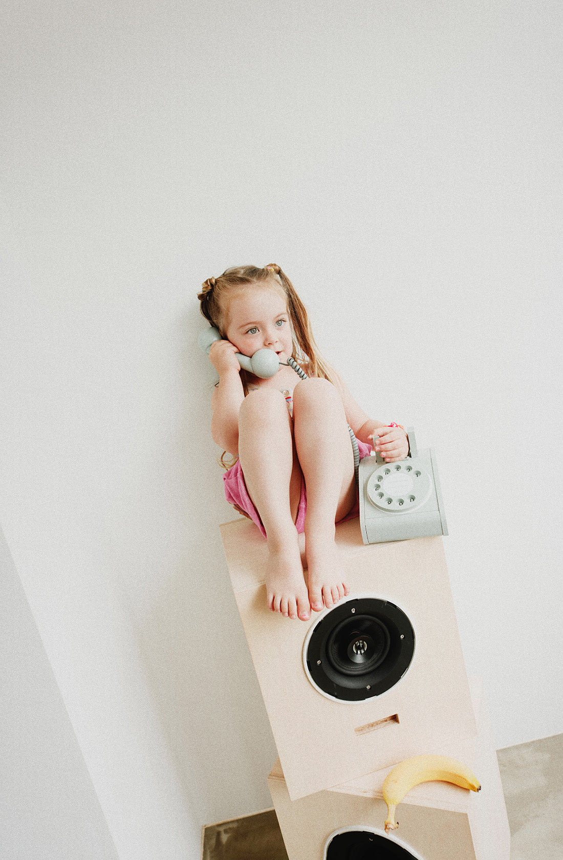 A young girl with blond hair styled in pigtails is sitting on top of a stack of speakers, holding a KIKO & GG Retro Telephone Green to her ear. She is barefoot, wearing a pink dress, and looking thoughtfully ahead. A banana is placed on one of the speakers below her, sparking children's imagination.