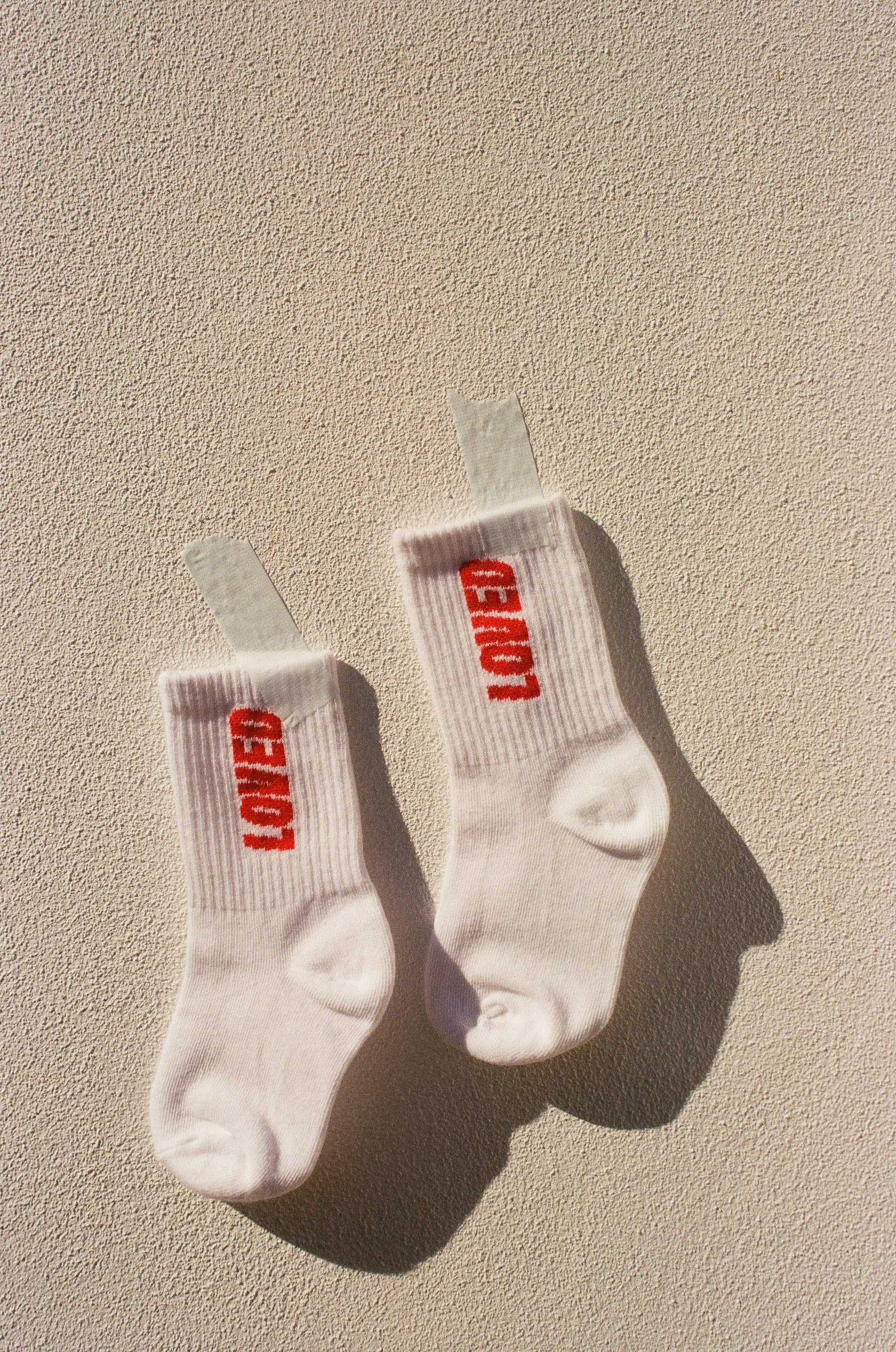 A pair of white Loved Sock baby sports socks from TINY LOVE CLUB, featuring the word "GENIUS" in bold red letters, are taped to a beige textured wall. The tape creates a shadow on the wall, while the elastic banded ankles ensure they stay snug for children aged 2-5 years.