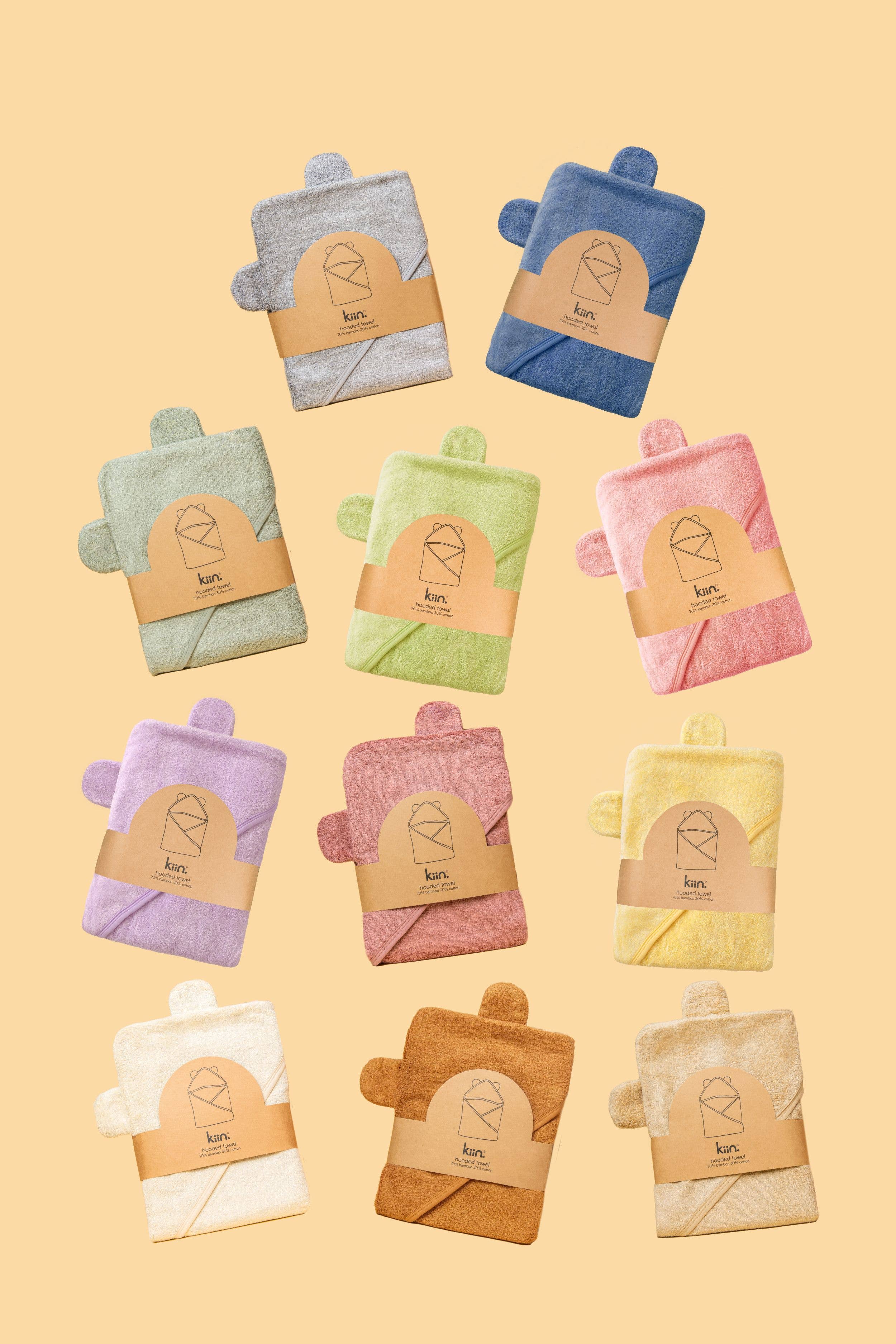 Ten vibrant washcloths in shades of gray, blue, green, yellow, pink, purple, and brown are set against beige. Made from hypo-allergenic bamboo fabric by KIIN BABY, each displays a cardboard label featuring a geometric logo with "Hooded Towel Ivory.