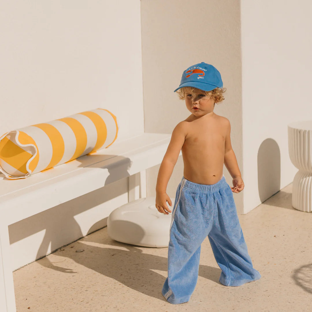 A curly-haired child stands shirtless in GOLDEN CHILDREN's Pool Blue Ciao Tutti Terry Towel Relaxed Pants, wearing a blue cap, positioned outdoors near a white bench and a yellow-striped cylindrical pillow.