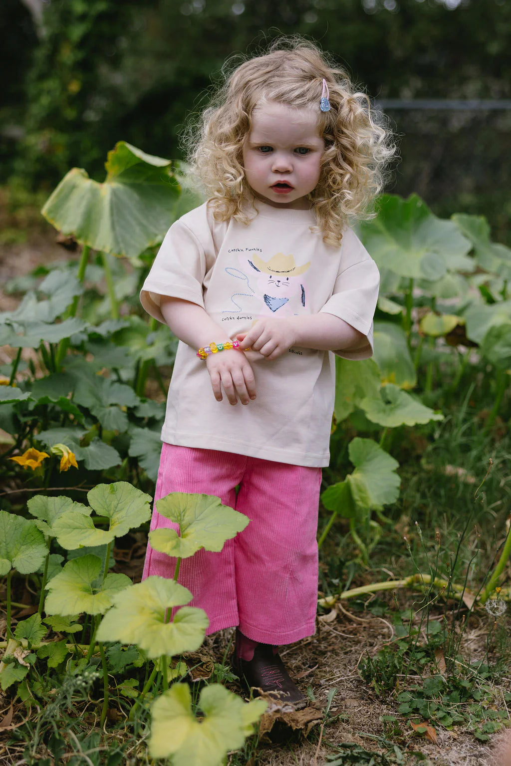 A young child with curly hair stands in a garden, wearing an oversized 100% cotton Sunday Siblings - Catch'n Bunnies Tee in pink. The beige T-shirt is adorned with playful illustrations and paired with pink pants and a beaded bracelet, surrounded by lush green plants.