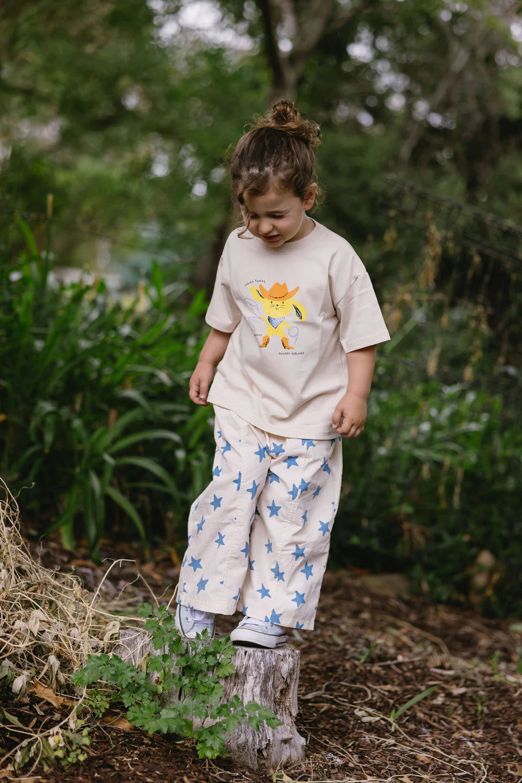 A young child with tied-up hair stands on a tree stump outdoors, wearing the Sunday Siblings - Catch'n Bunnies Tee Yellow by SUNDAY SIBLINGS and blue star-patterned pants, surrounded by green plants and trees.
