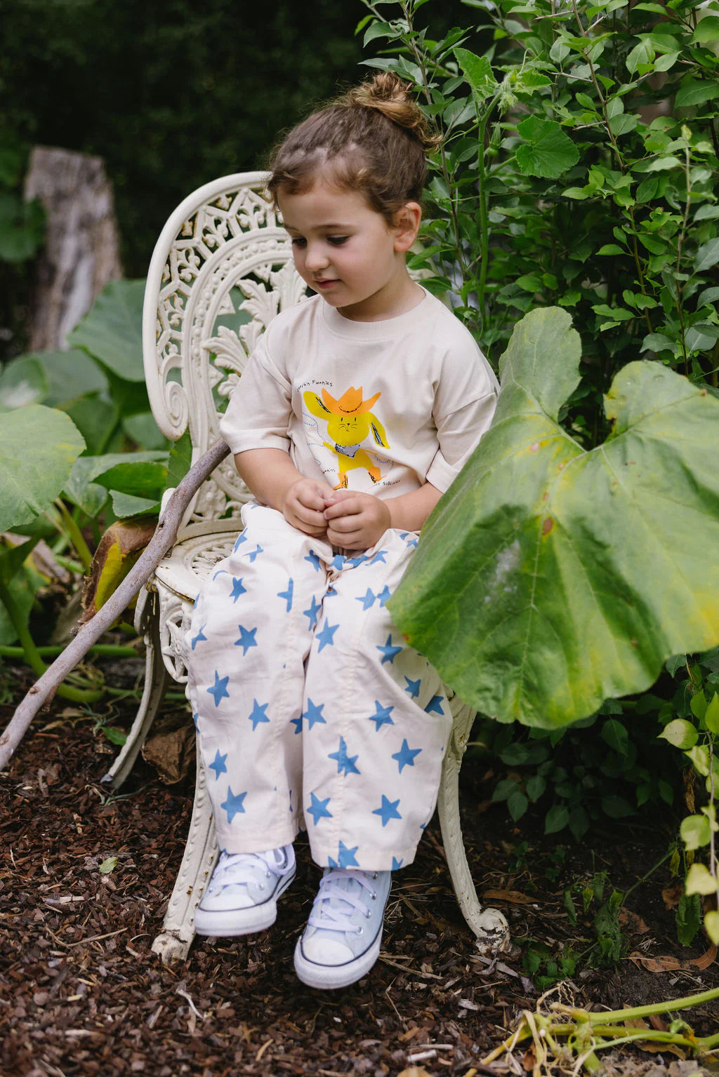 A young child with brown hair in a bun sits on a white garden chair, wearing the SUNDAY SIBLINGS Sunday Siblings - Catch'n Bunnies Tee Yellow and white pants with blue stars, surrounded by lush green leaves in a garden.