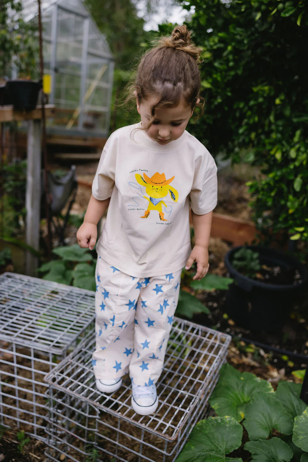 A young child with brown hair in a bun stands on a metal grid outdoors, wearing the SUNDAY SIBLINGS Sunday Siblings - Catch'n Bunnies Tee Yellow, surrounded by plants and greenery.