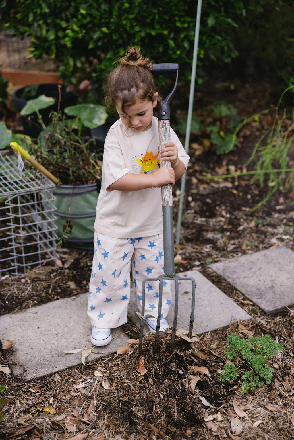 A young child in the SUNDAY SIBLINGS Catch'n Bunnies Tee Yellow stands in a garden holding a pitchfork, surrounded by leaves, plants, and gardening tools.