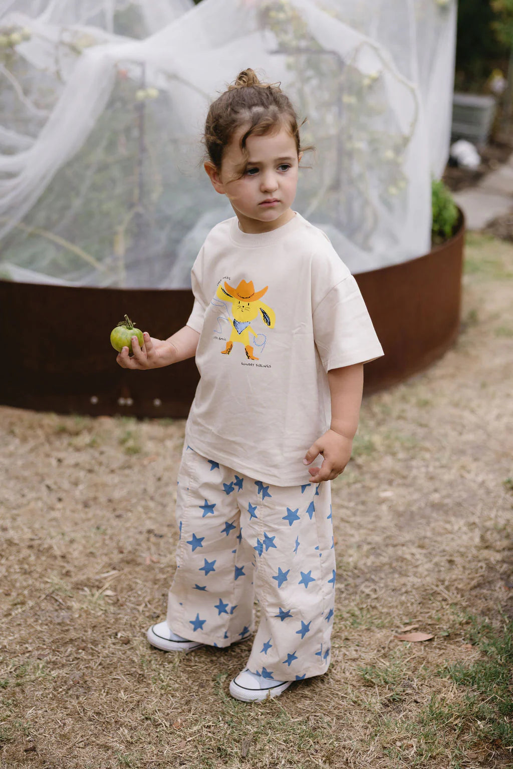 A young child stands on grass holding a green fruit, wearing the SUNDAY SIBLINGS Catch'n Bunnies Tee Yellow—an oversized 100% cotton cream t-shirt with a cartoon print—and wide cream pants with blue stars. A garden netted structure is visible in the background.
