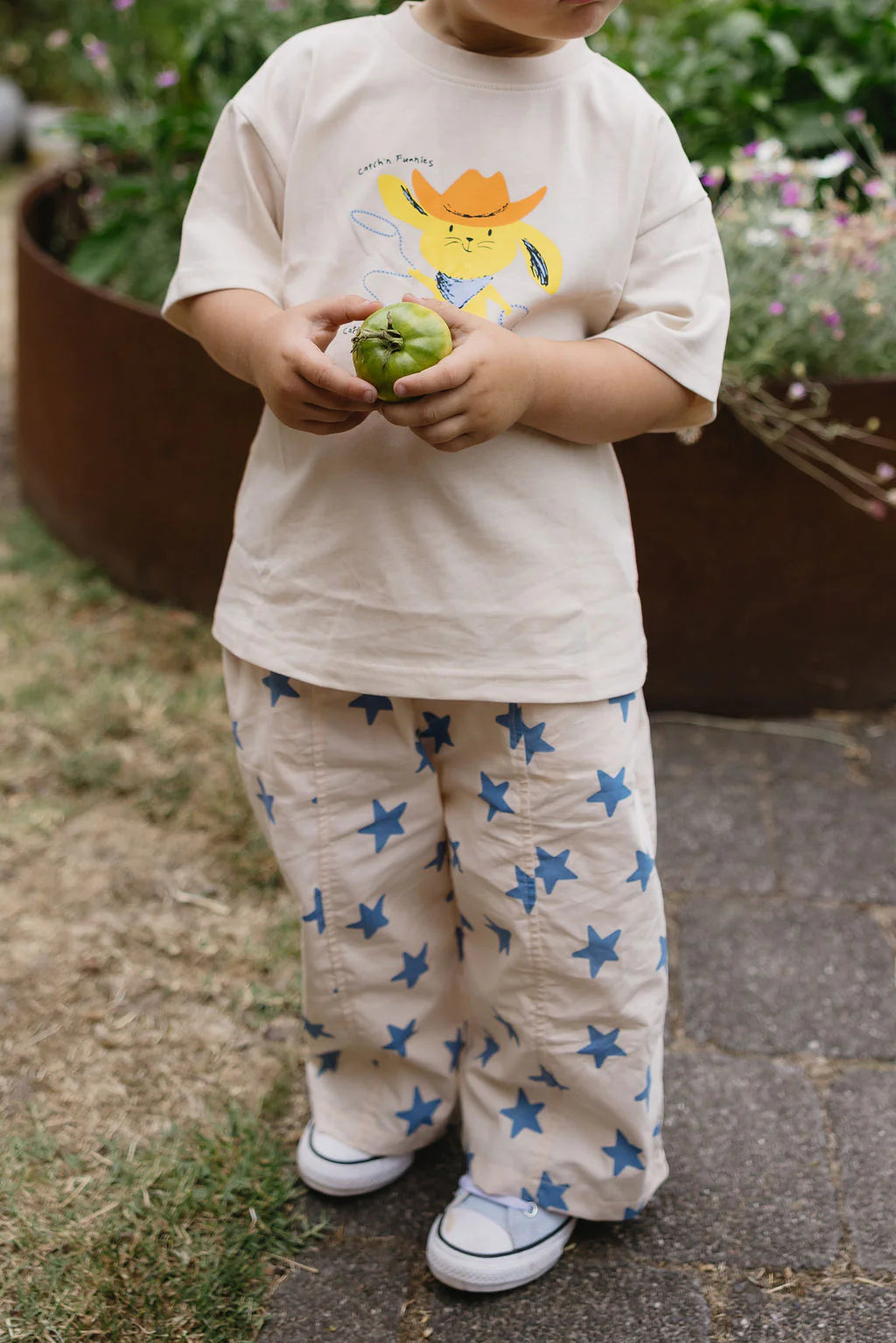 A child stands outdoors holding a green fruit, wearing the SUNDAY SIBLINGS Catch'n Bunnies Tee in yellow and beige pants with blue stars. They pair this with white sneakers amidst grass, flowers, and a circular planter.