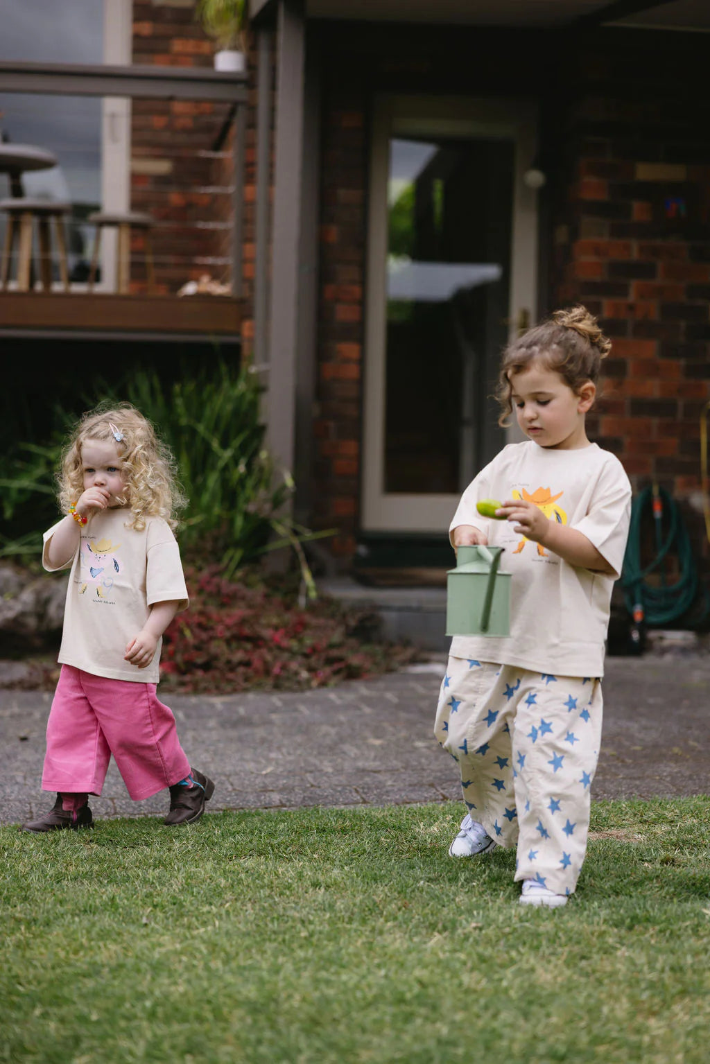 Two young children play outside on grass. One wears the SUNDAY SIBLINGS Catch'n Bunnies Tee in yellow with pink pants, holding a flower. The other sports star-patterned pants and carries a green watering can. A brick house and garden are behind them.