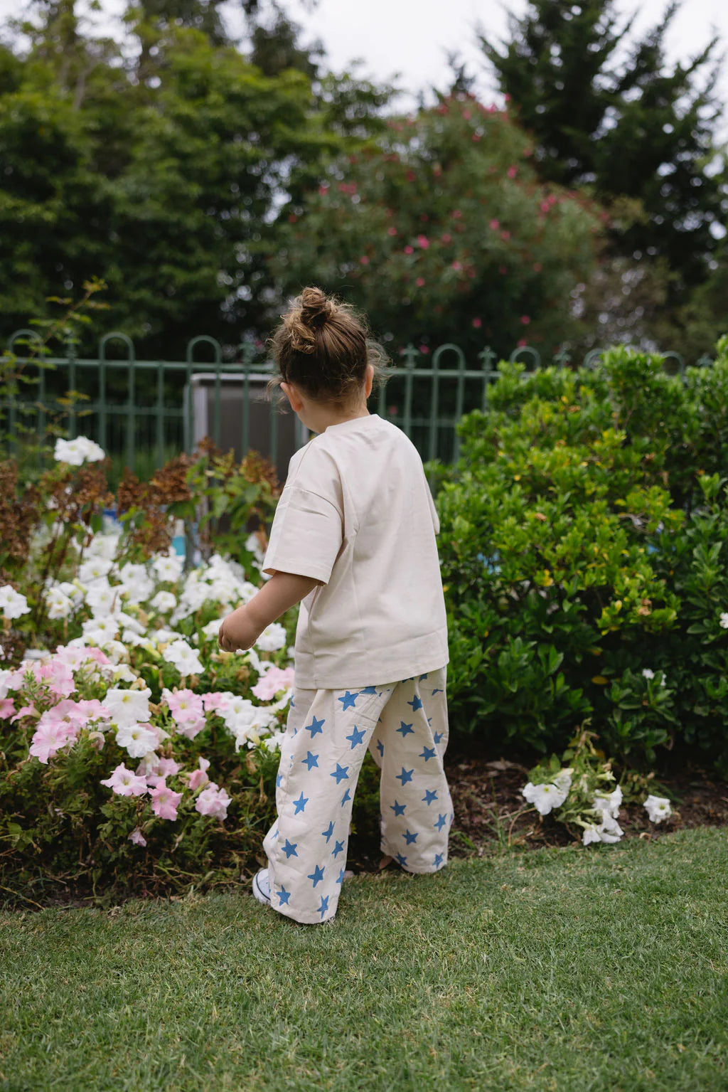 A young child with hair in a bun, wearing the SUNDAY SIBLINGS Sunday Siblings - Catch'n Bunnies Tee Yellow, stands on grass outside, looking at blooming flowers and green bushes in a garden.