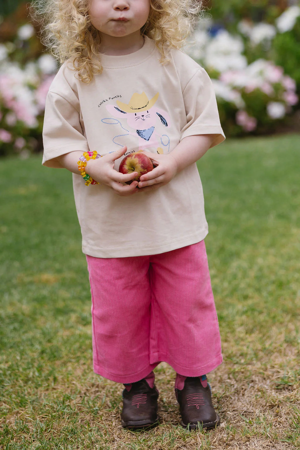 A young curly-haired blonde child stands on grass, holding a peach. They wear a SUNDAY SIBLINGS Catch'n Bunnies Tee in pink, bright pink pants, brown shoes, and a colorful bracelet. Flowers add vibrancy to the background.