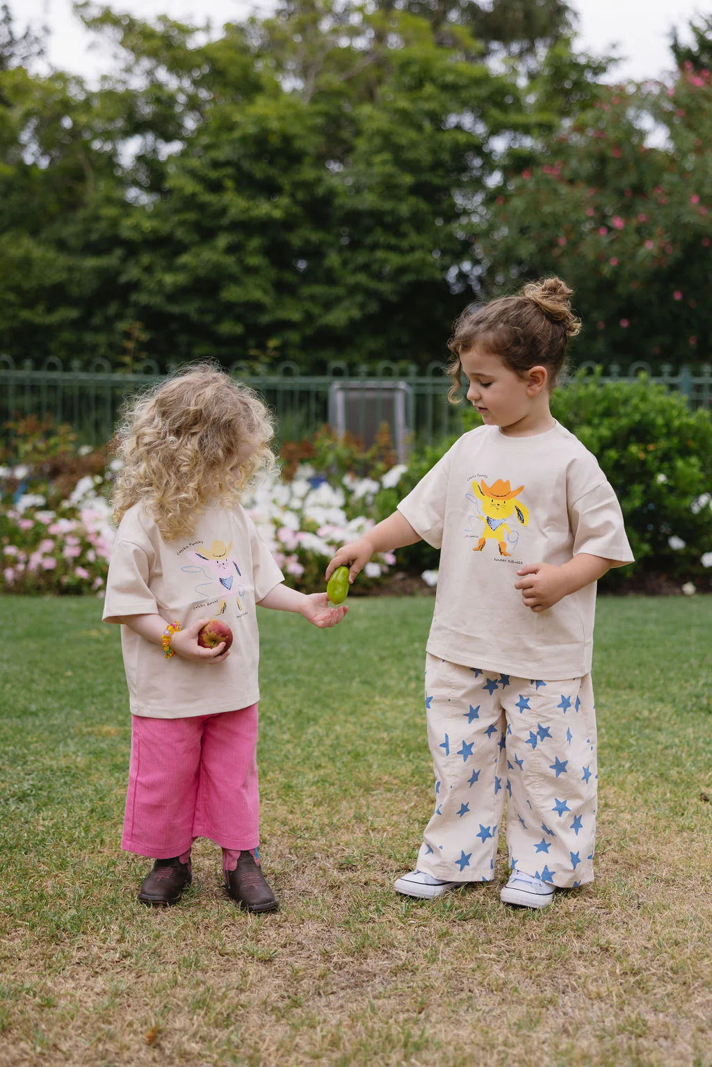 Two young children play on grass, one in pink pants and brown boots, the other in star-patterned pants. They wear the SUNDAY SIBLINGS Catch'n Bunnies Tee Yellow, a 100% cotton oversized Easter shirt, exchanging a green object and holding apples.