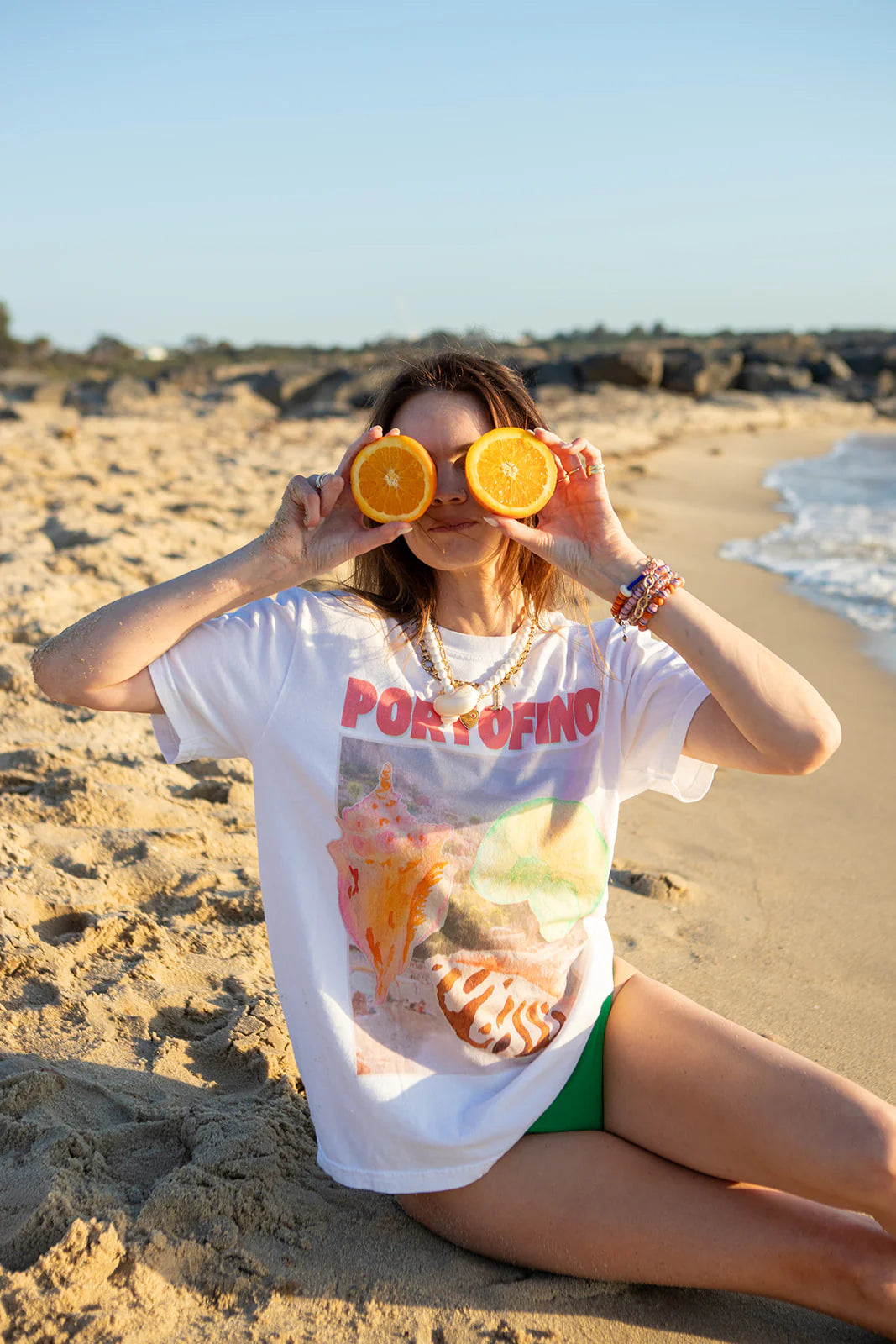 On a sandy beach, someone playfully holds orange halves over their eyes while wearing the MAKU THE LABEL's Portofino T-Shirt, made of 100% cotton, paired with green shorts. The clear sky and gentle waves provide the perfect backdrop for this whimsical scene.
