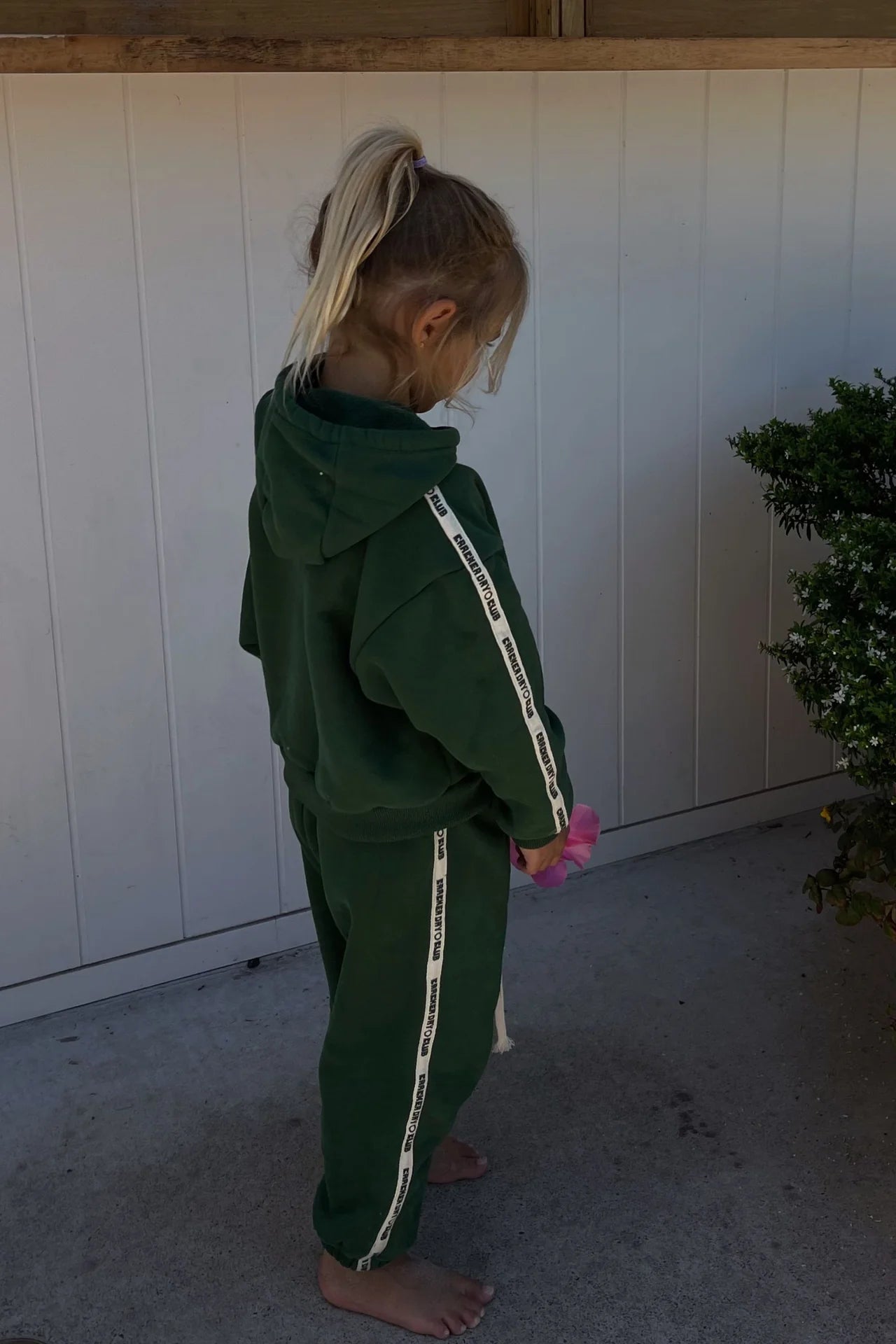 A young child with blonde hair in a ponytail stands barefoot outdoors near a white fence, wearing CRACKER DAY CLUB’s Cracker Day Club - Kids Cloud Racer Sweatpants Forest, featuring white striped text on the legs.
