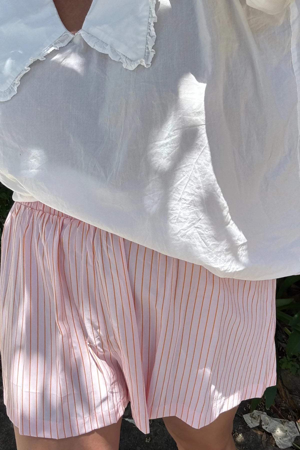 A person in a large-collared white shirt and Mama Everyday Shorts Sunset Stripe by LITTLE THE LABEL stands outdoors. Sunlight casts textured shadows on the outfit, with partial green foliage in the background, embodying an effortless Mama Styles vibe.